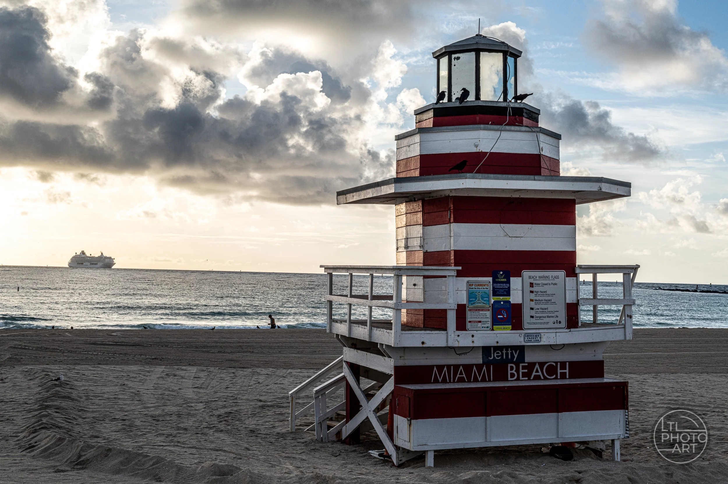 Red-and-white Jetty lifeguard tower on South Beach in Miami, Florida, with dramatic clouds, the Atlantic Ocean, and a cruise ship in the distance.