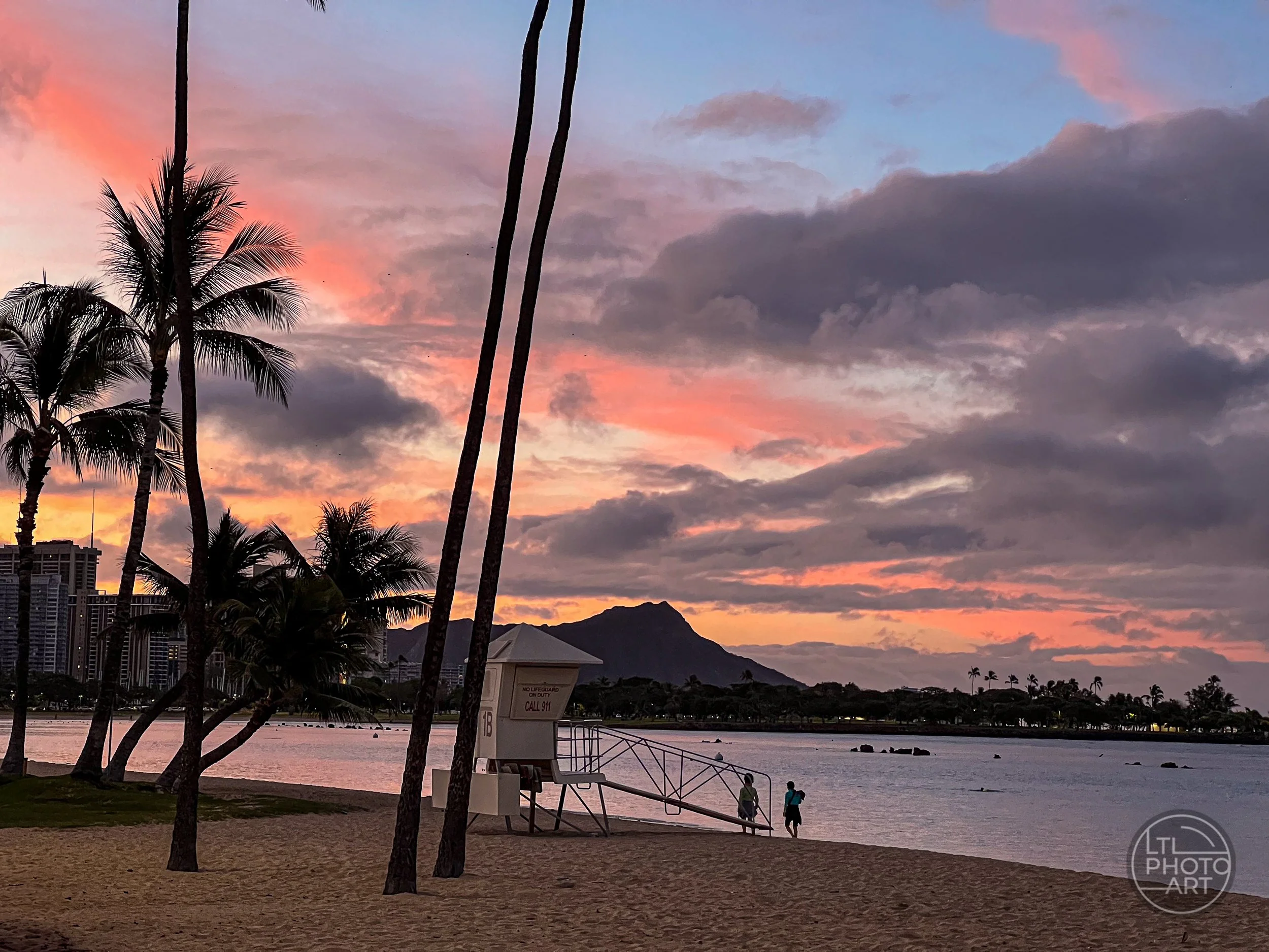 Lifeguard tower at Ala Moana Regional Park in Honolulu, Oahu, at sunrise with palm trees, colorful clouds, and Diamond Head in the background.