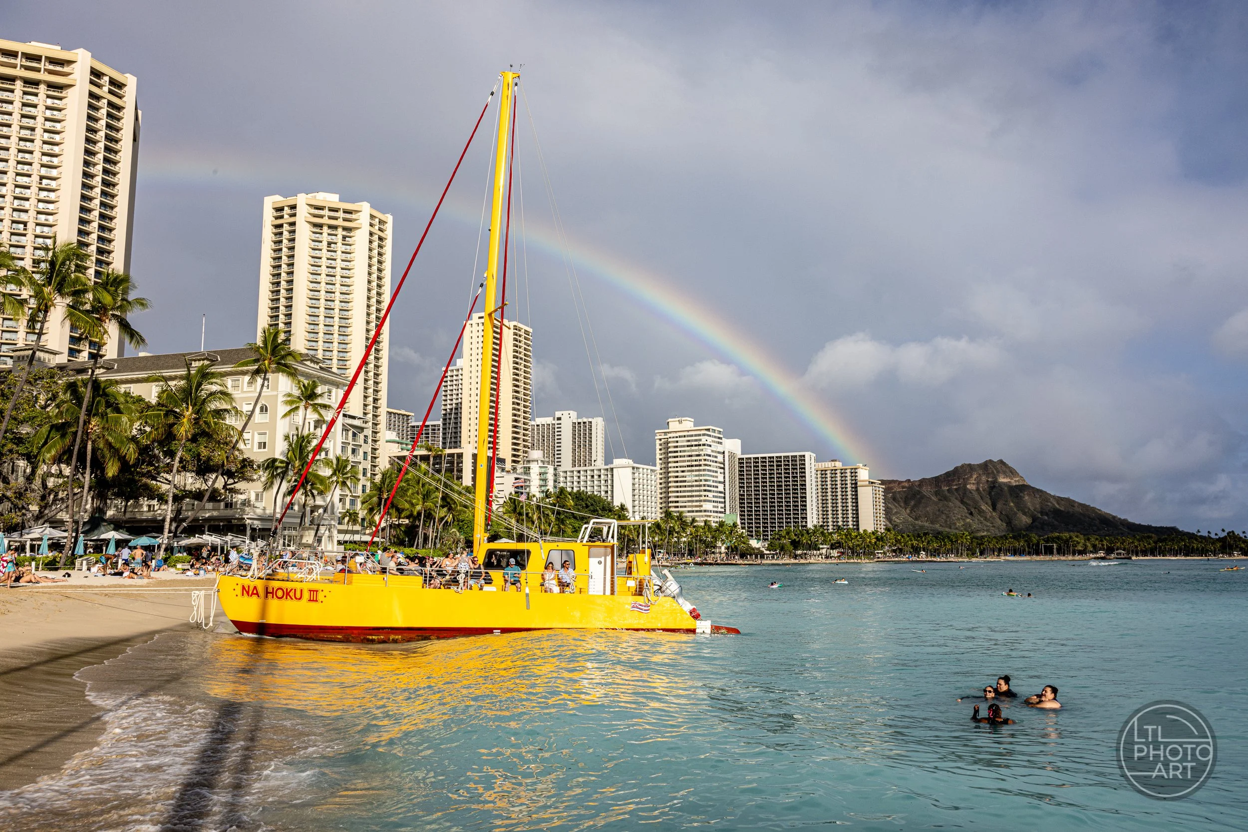 The yellow Na Hoku III catamaran at Waikiki Beach on Oahu, with swimmers in the water, a rainbow in the sky, and Diamond Head in the background.