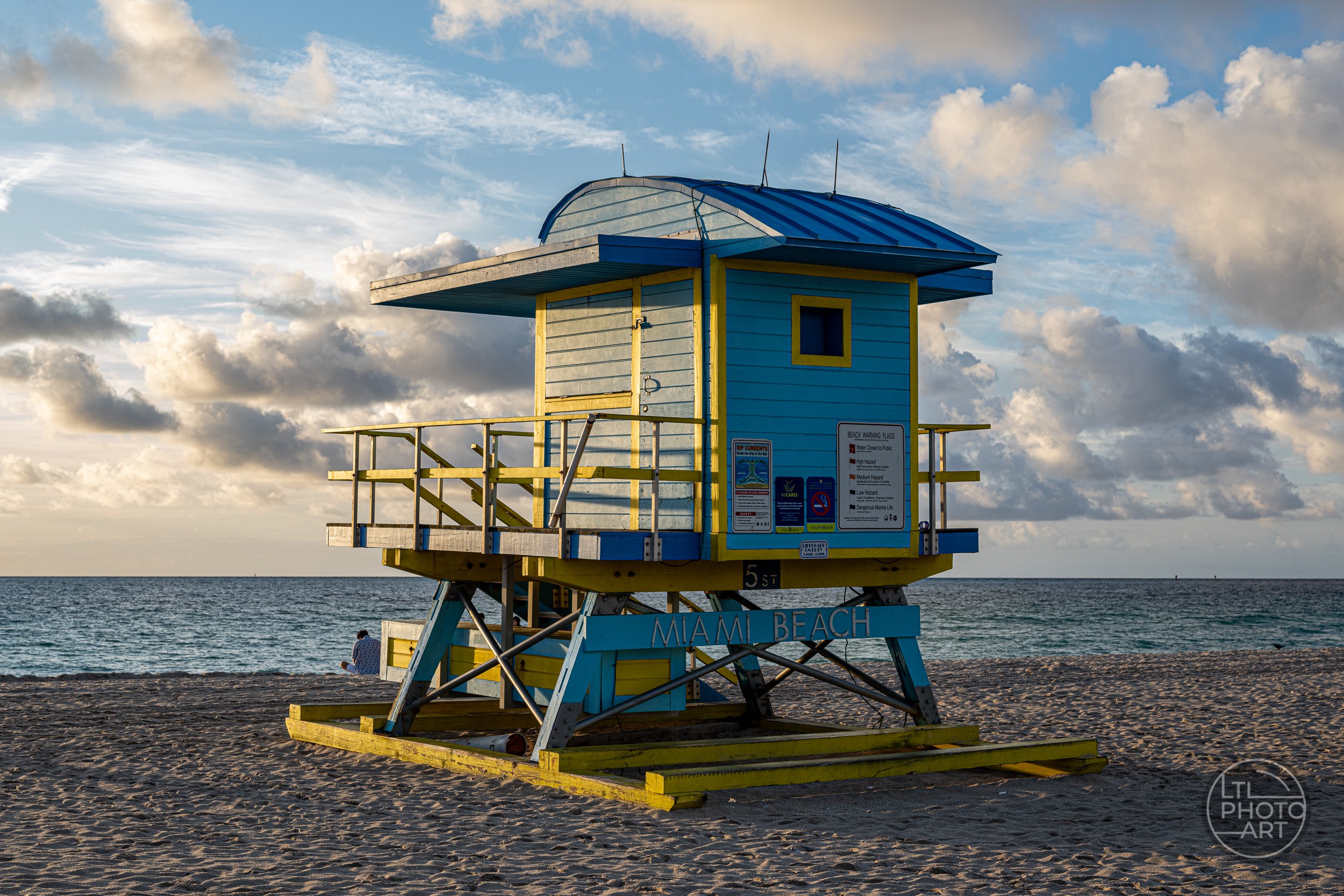 Blue and yellow 5th Street lifeguard tower on South Beach in Miami, Florida, with the Atlantic Ocean and clouds in the background.