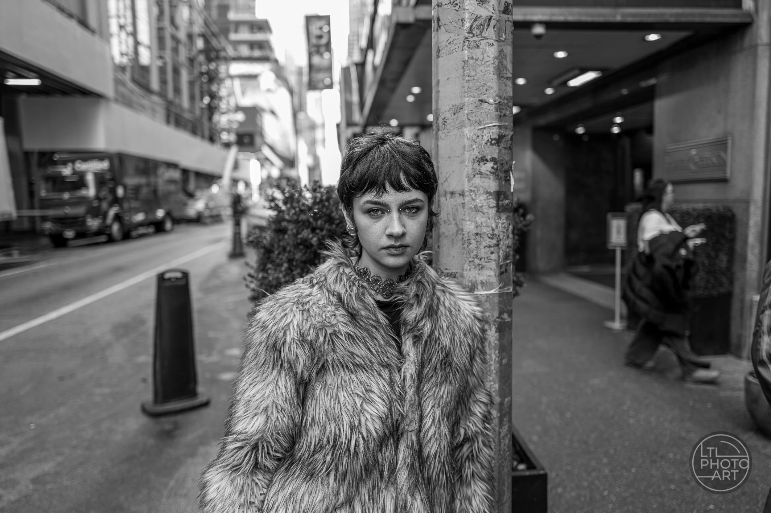 Black-and-white street portrait of a young woman wearing a textured coat beside a utility pole in New York City.