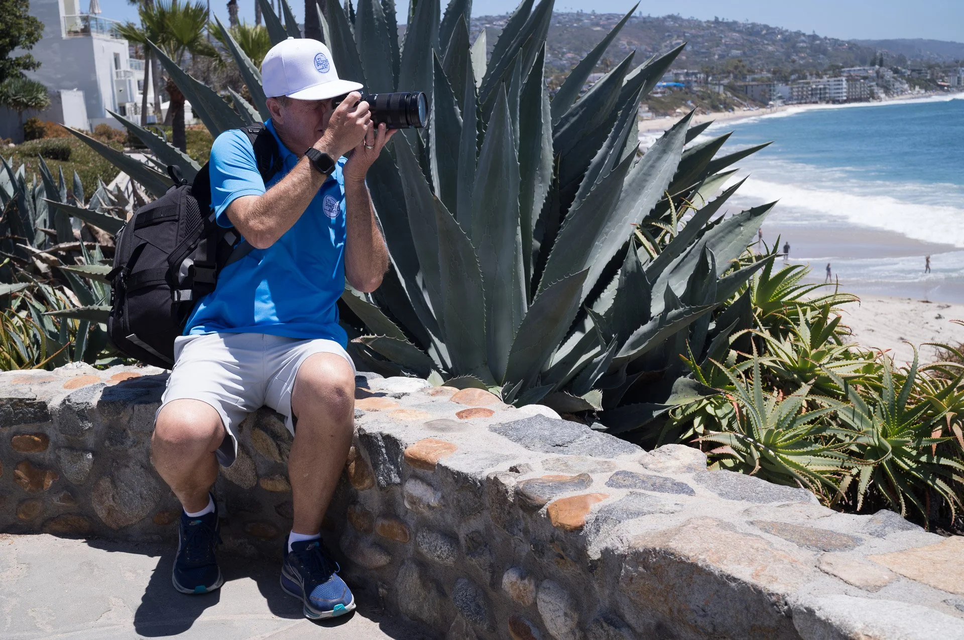 Photographer Lyle Lansdell using a camera beside large coastal plants with Laguna Beach and the Pacific Ocean in the background.
