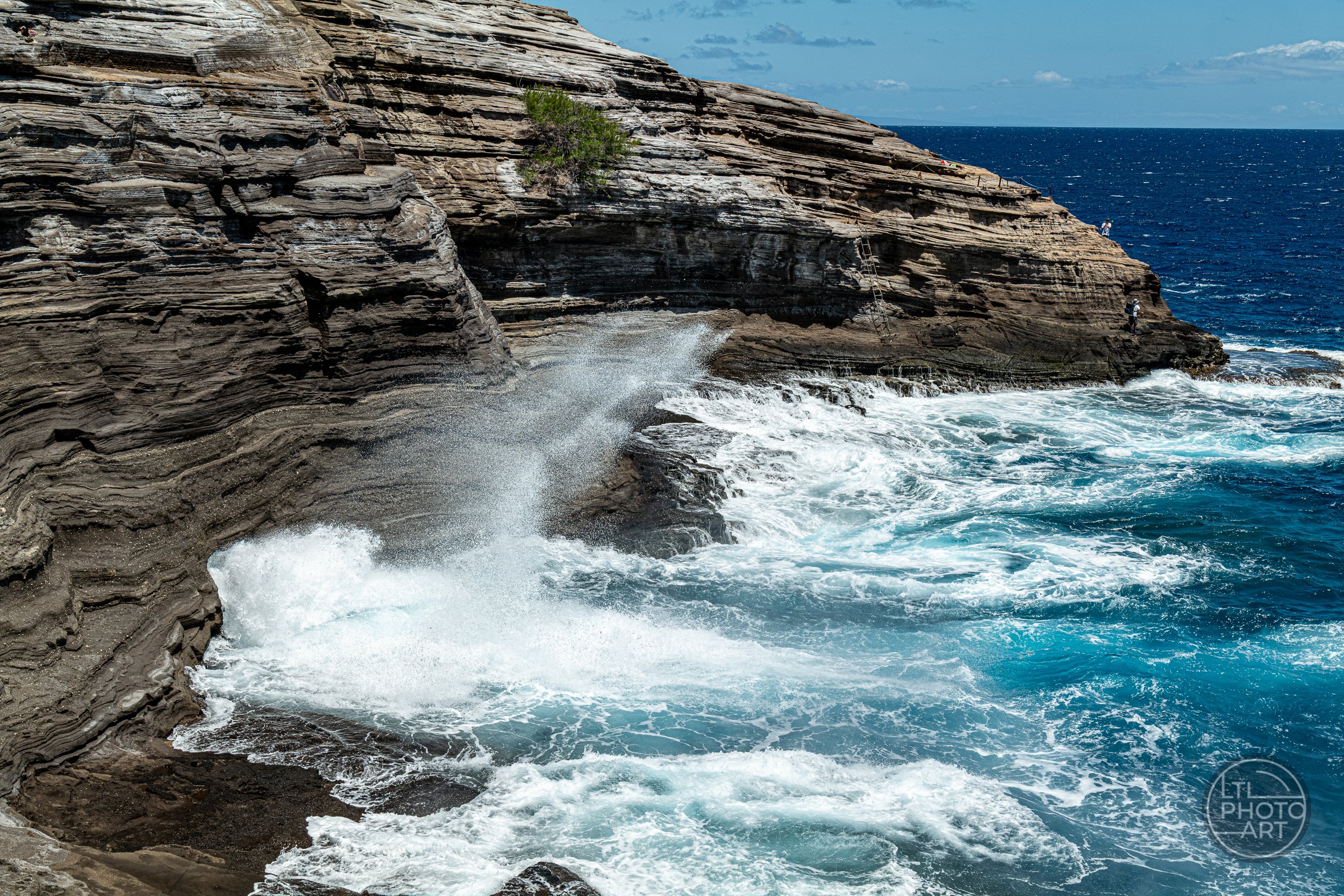 Ocean spray bursting from Spitting Cave on Oahu, Hawaii, with turquoise water, layered rock cliffs, and small fishermen visible on the distant shoreline.