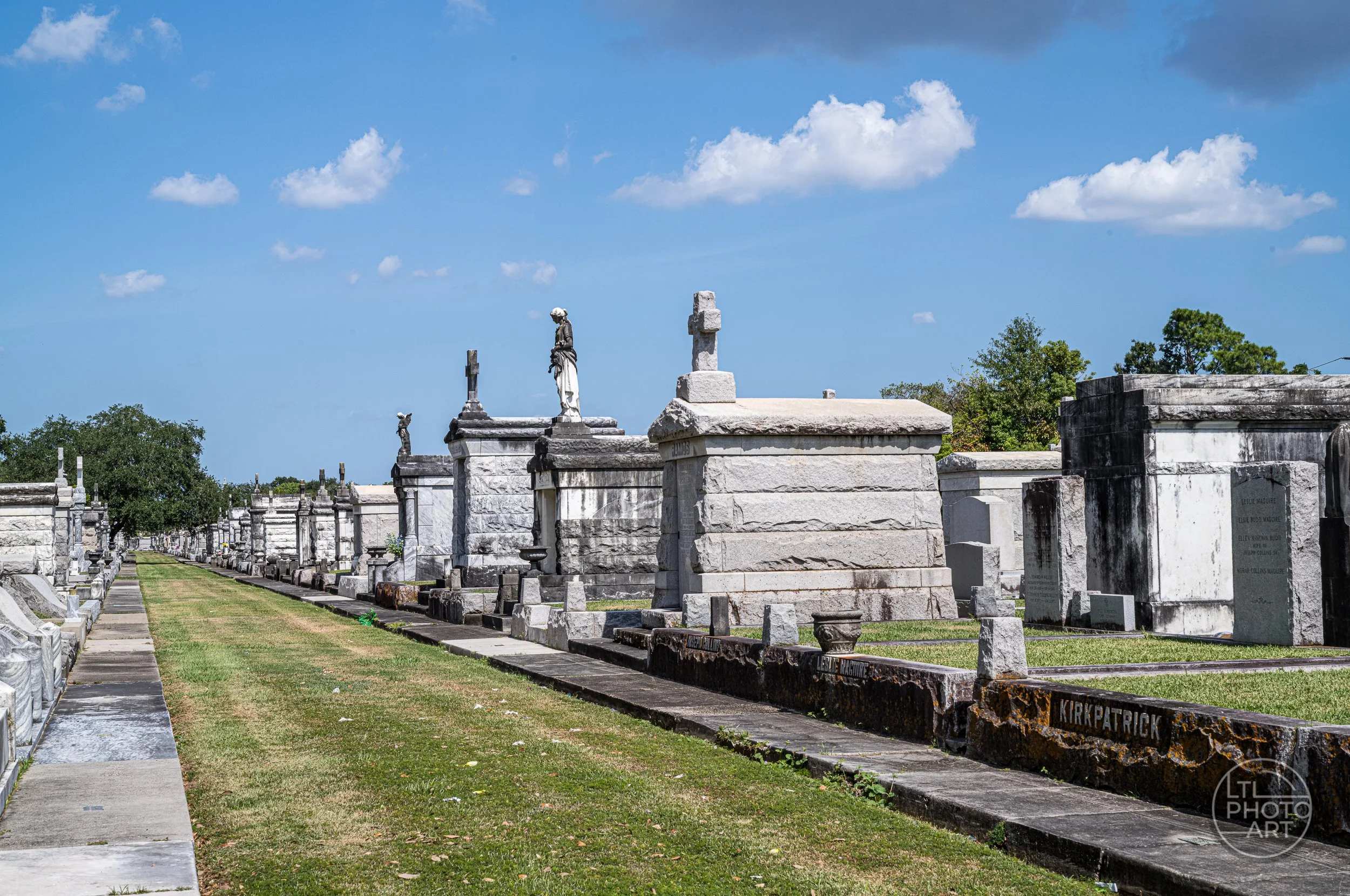 Rows of above-ground tombs in Metairie Cemetery in New Orleans, with statues, stone monuments, grass paths, and blue sky.