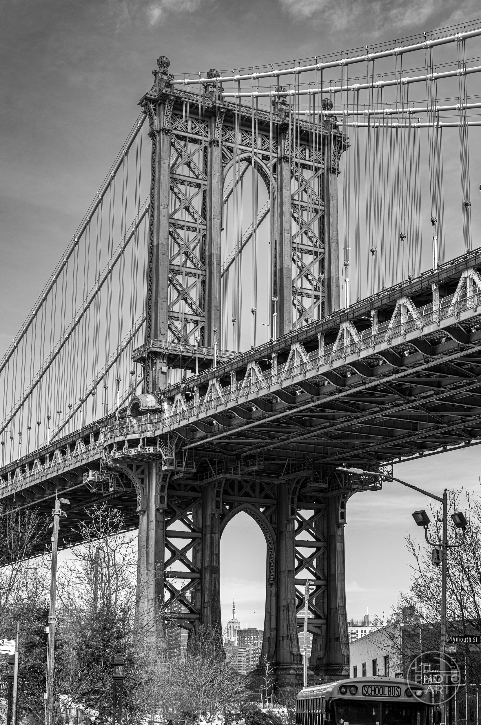 Black-and-white street view of the Manhattan Bridge from Washington Street in DUMBO, Brooklyn, with pedestrians, parked cars, and a school bus.