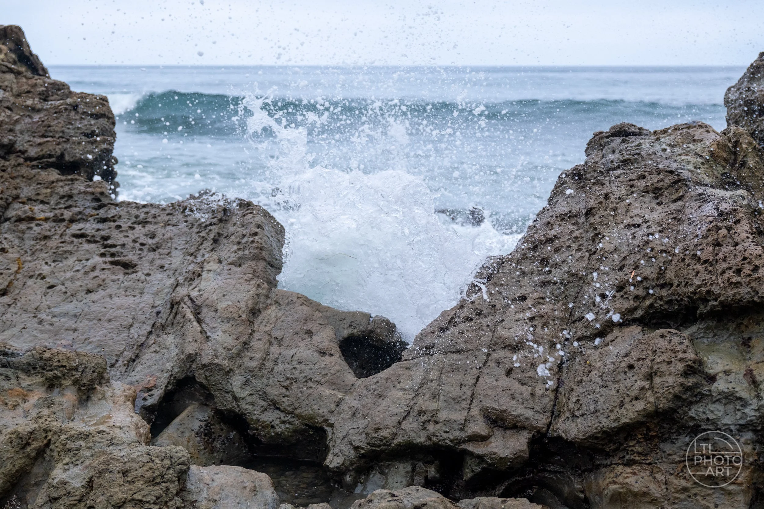 Wave crashing through rocky tide pools near Rockpile Beach in Laguna Beach, with white spray rising between the foreground rocks and the Pacific Ocean beyond.