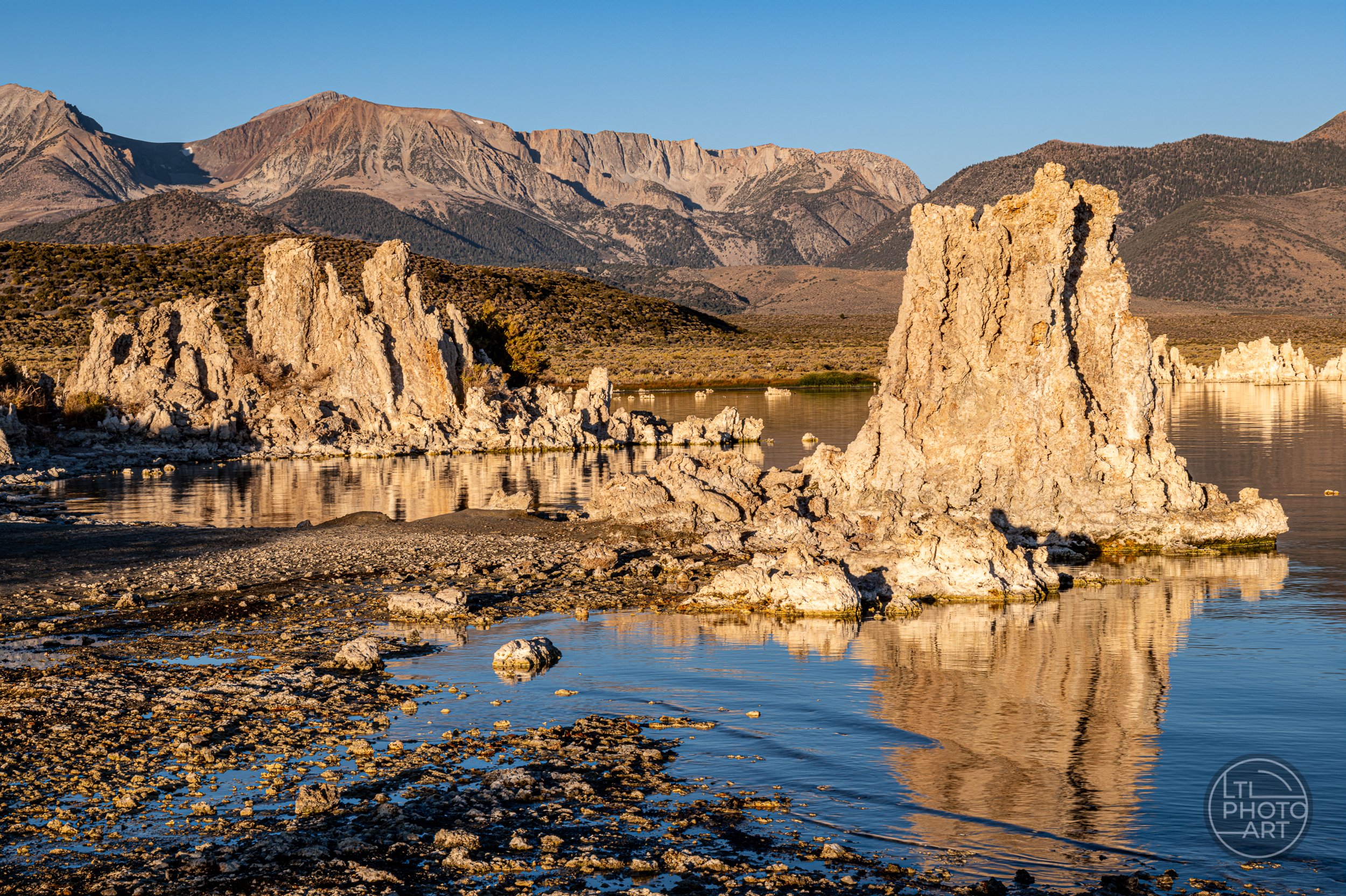 Landscape photograph of limestone tufa towers at Mono Lake, California, illuminated by warm golden sunlight, with reflections in the calm water and rugged Eastern Sierra mountains in the background.