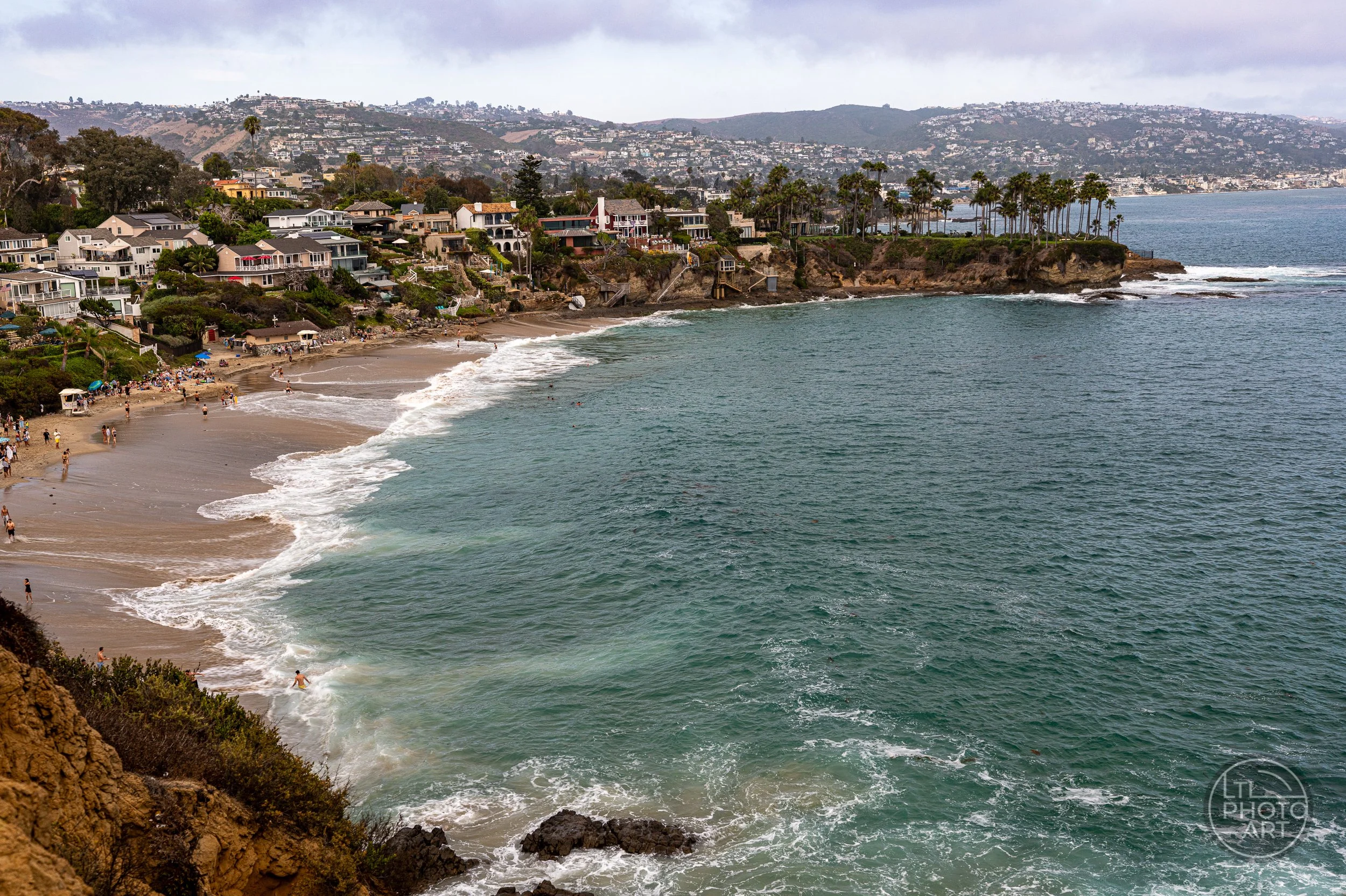 Elevated view of Crescent Bay with beachgoers, coastal homes, palm trees, and turquoise water in Laguna Beach, California.