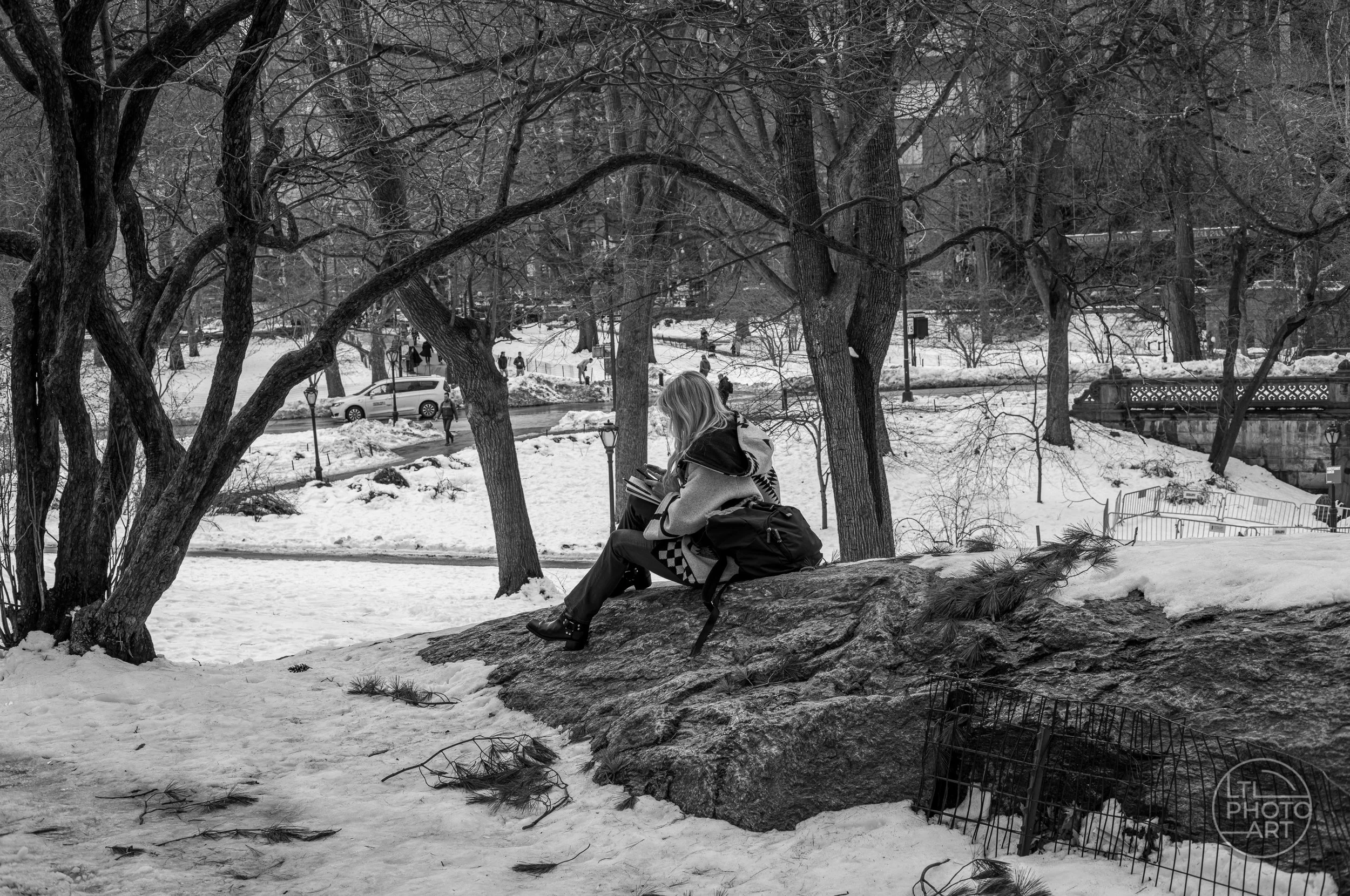 Black-and-white photograph of a woman using her phone while seated on a rock in snow-covered Central Park, New York City.