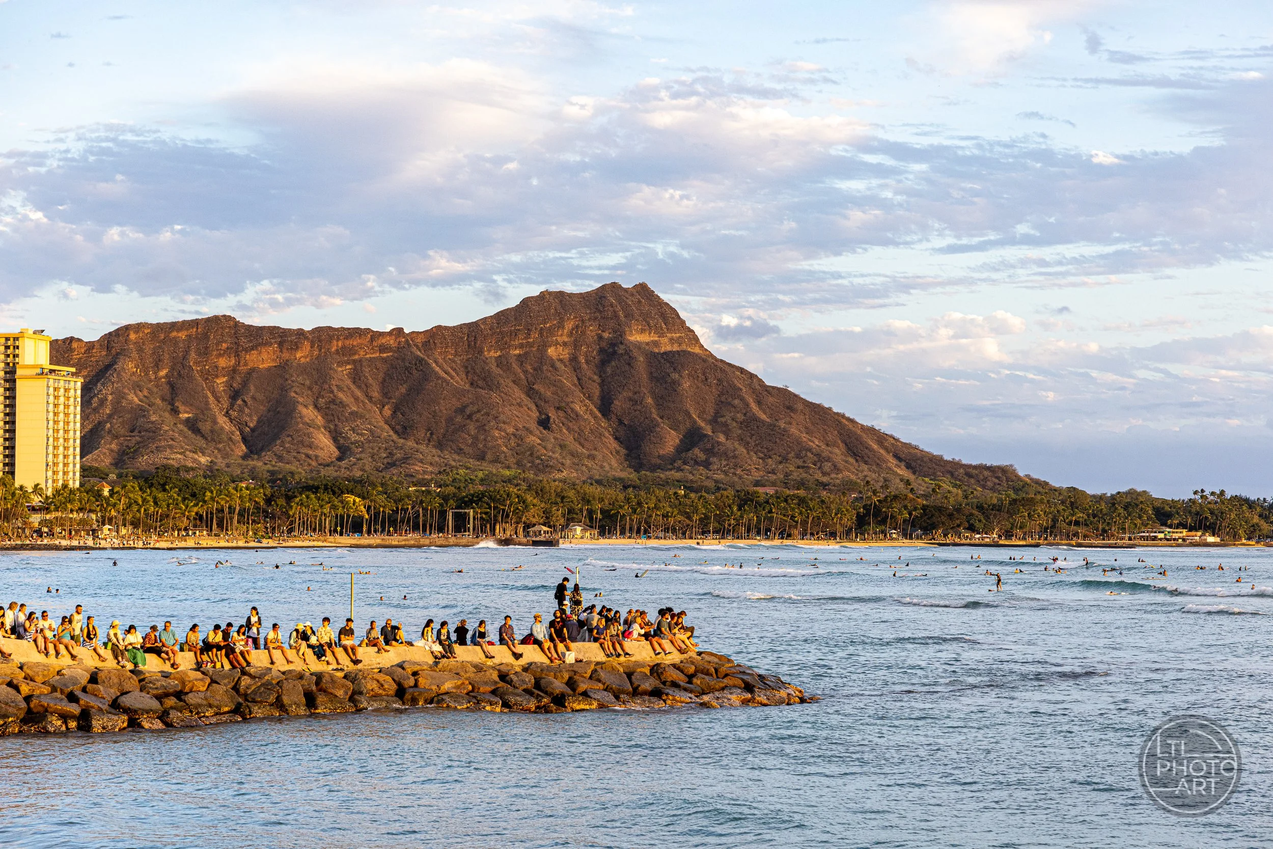 Diamond Head illuminated by warm sunset light beyond Waikiki Beach, with people gathered on a rocky point and surfers in the water off Oahu, Hawaii.