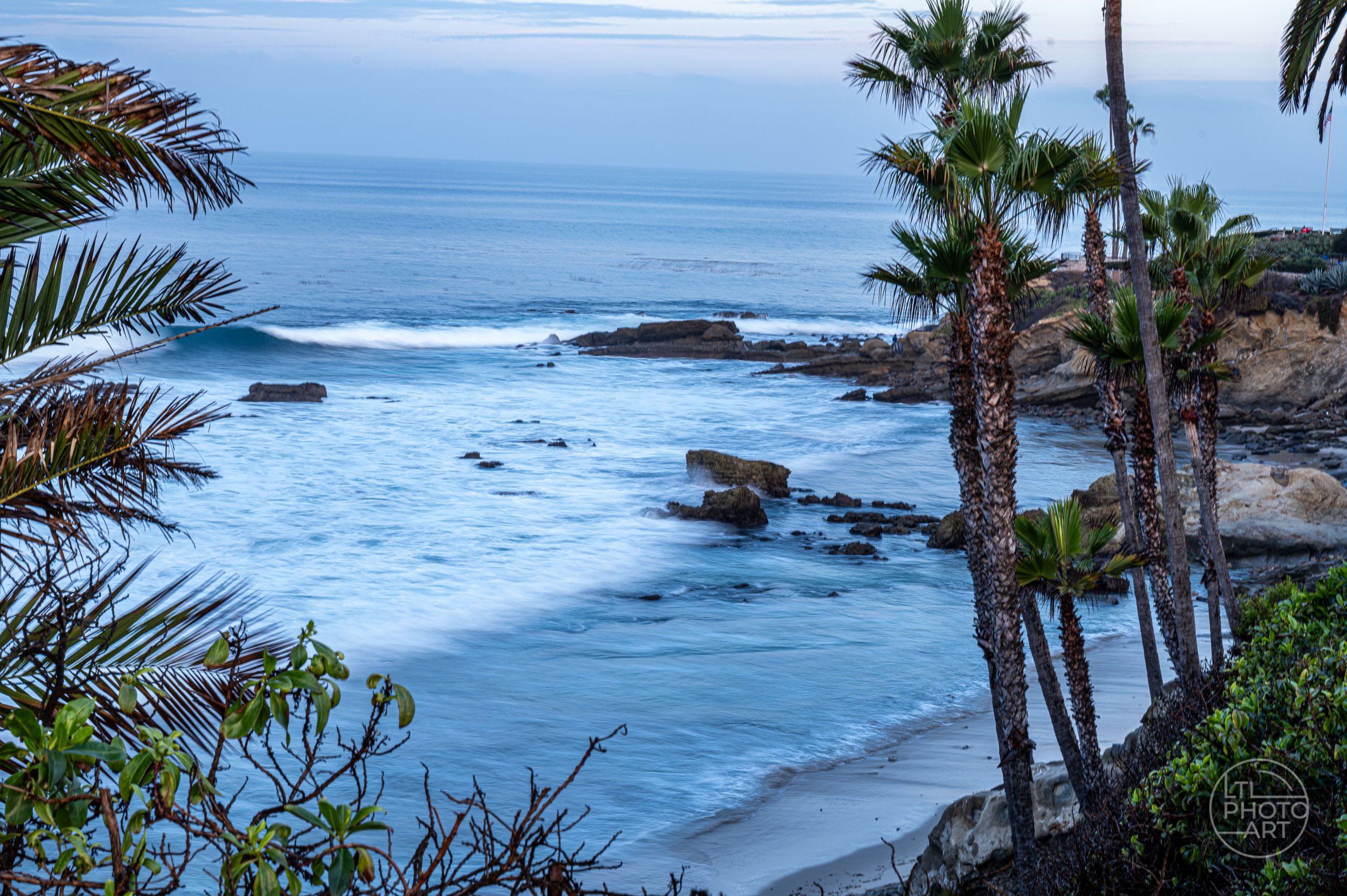 Slow-shutter coastal view of Monument Point framed by palm trees from an elevated path in Heisler Park, Laguna Beach.