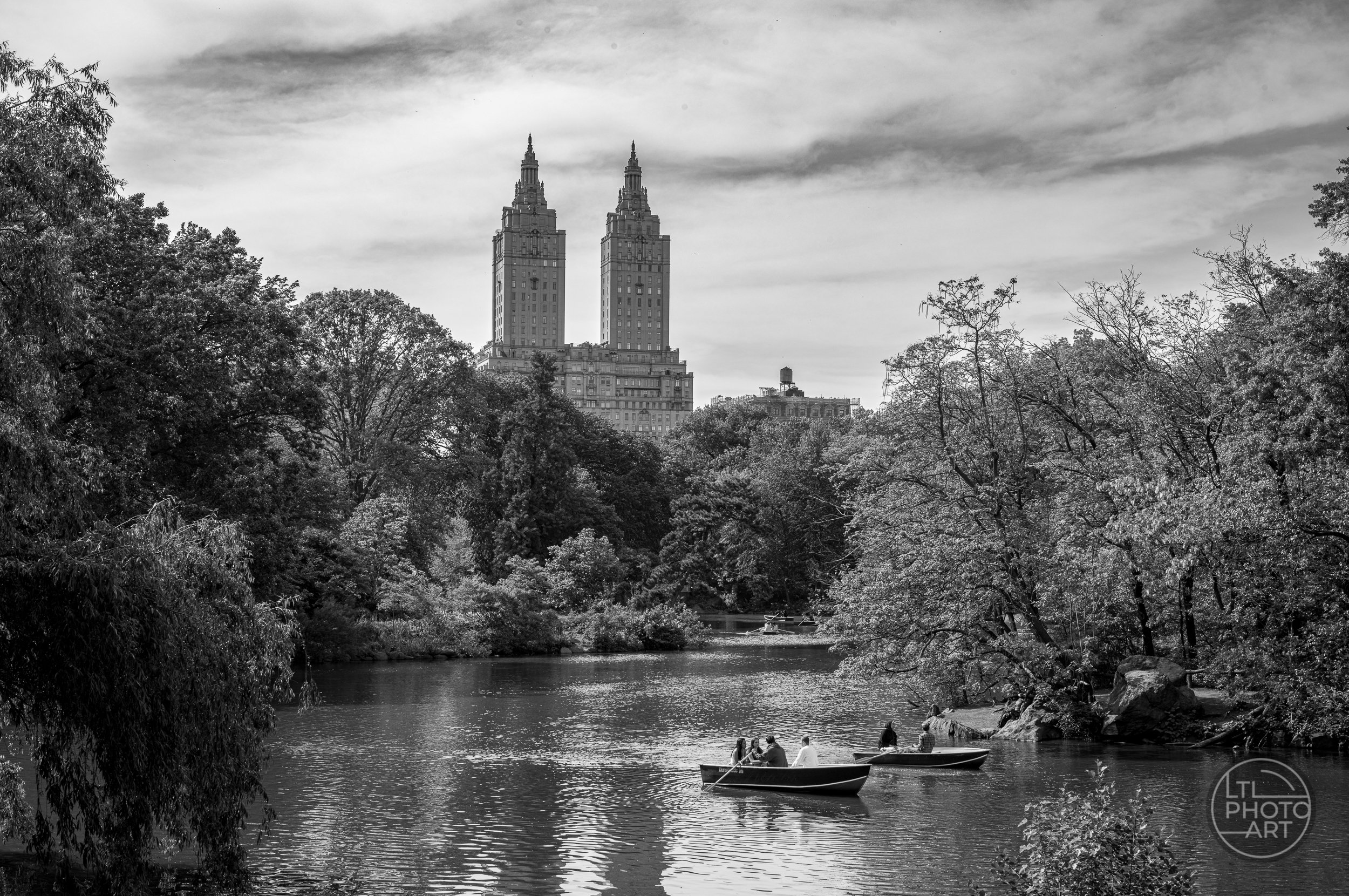 Black-and-white view of rowboats on The Lake in Central Park, with trees and the twin towers of the San Remo in the background.