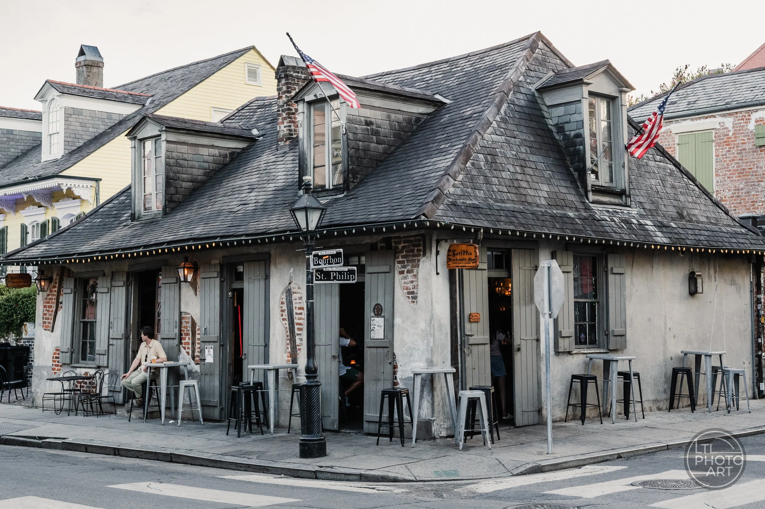 Lafitte’s Blacksmith Shop in New Orleans, with historic weathered architecture, flags, street seating, and a person waiting outside.