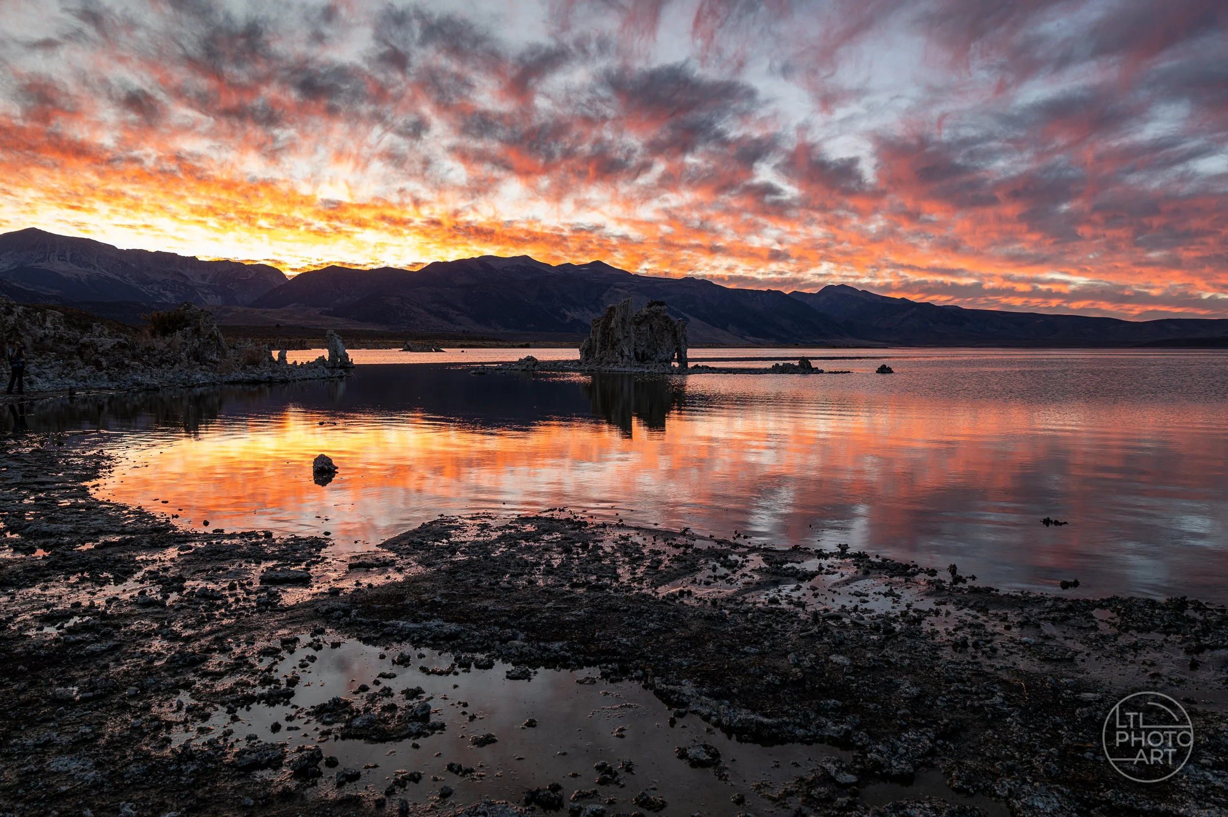 Sunset at Mono Lake, California, with orange and pink clouds reflected in the water, distinctive limestone tufa formations rising from the lake, and the Eastern Sierra mountains silhouetted along the horizon.