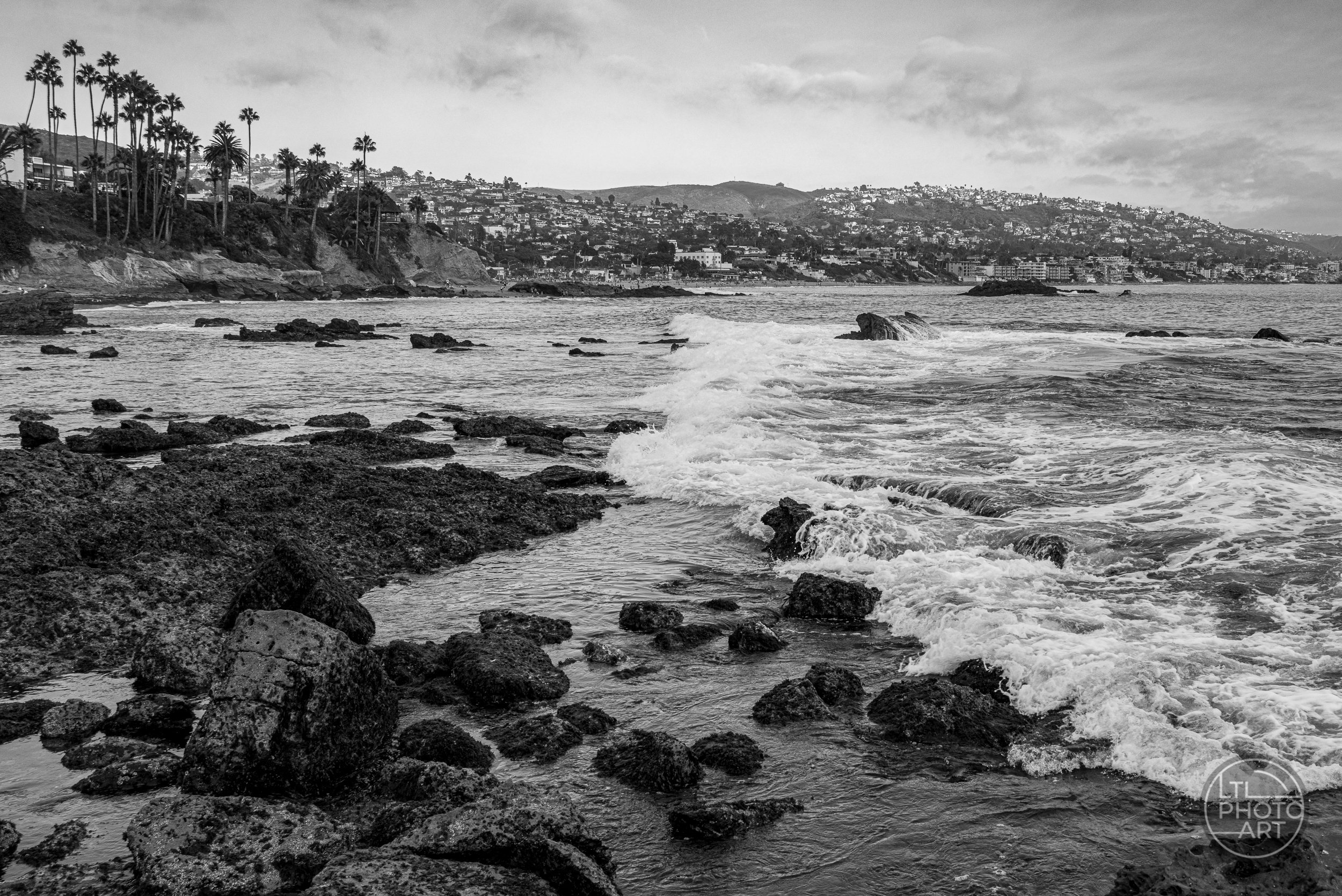 Black-and-white photograph of waves rolling across the rocky shoreline at Monument Point toward Rockpile Beach, with Laguna Beach hillsides in the background.