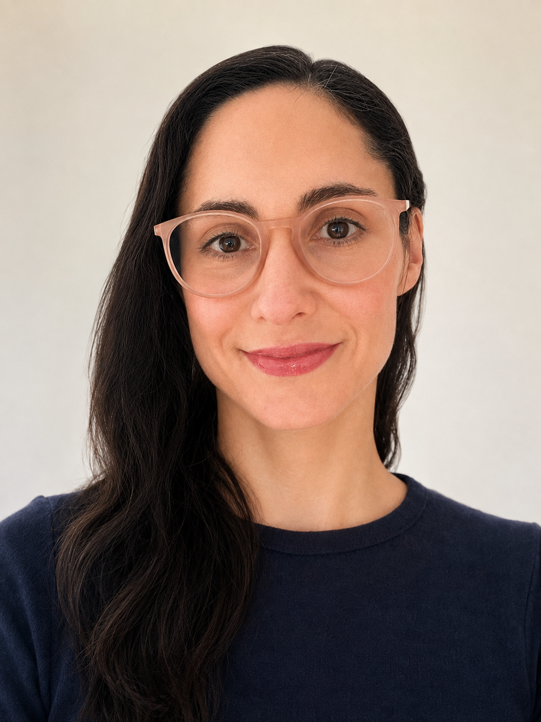 A woman with long dark hair, wearing pink glasses and a navy blue shirt, is smiling and looking at the camera against a plain light background.