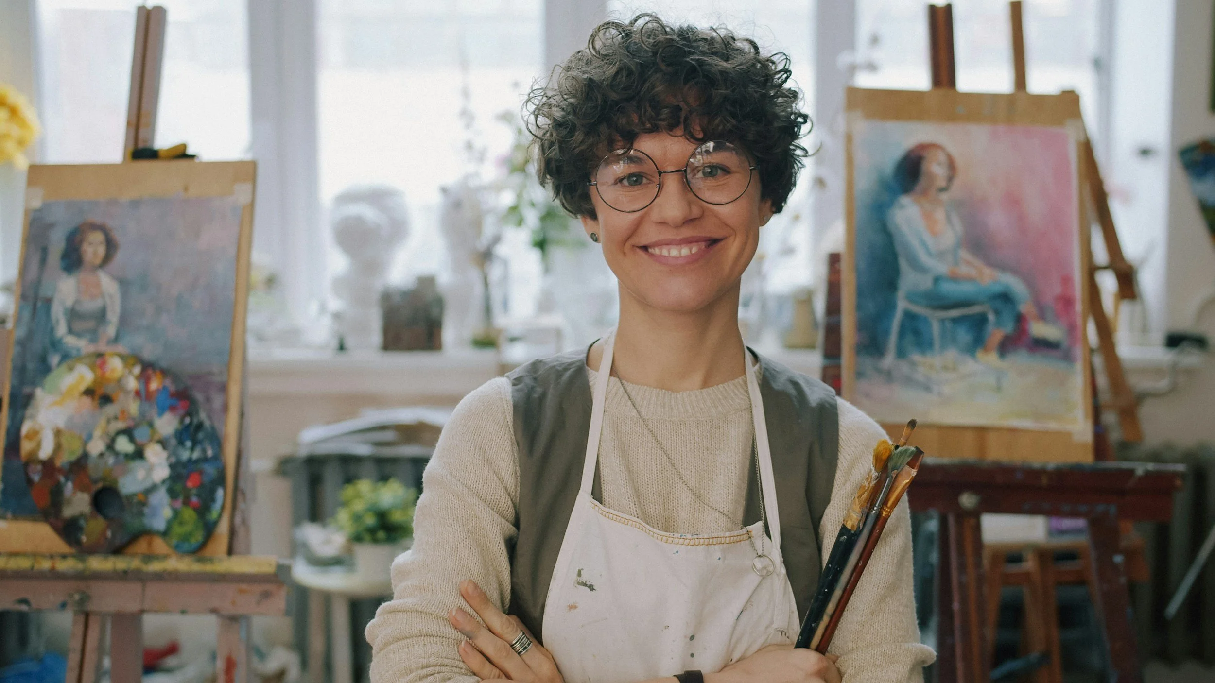 Smiling creative woman with curly hair and glasses standing in an art studio, surrounded by paintings and art supplies.