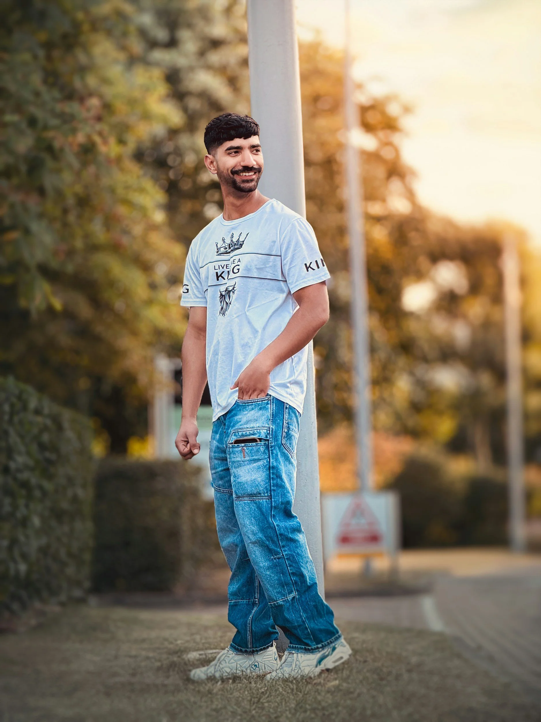 A young man with dark hair and a beard wearing a white graphic t-shirt and blue jeans standing outdoors during sunset, smiling and looking to his left.