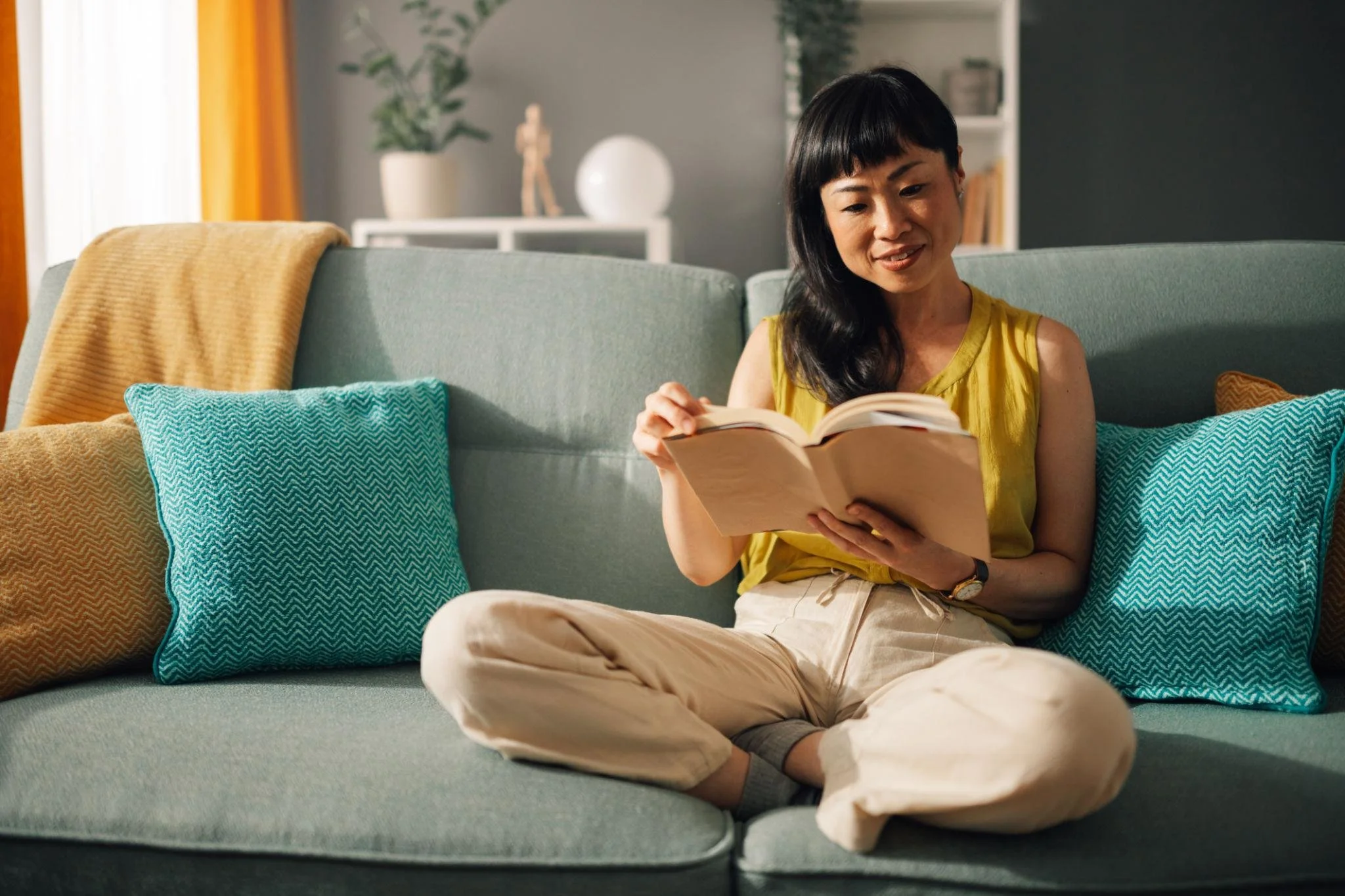Woman sitting cross-legged on a sofa, reading a book, with colorful pillows around her in a cozy living room.