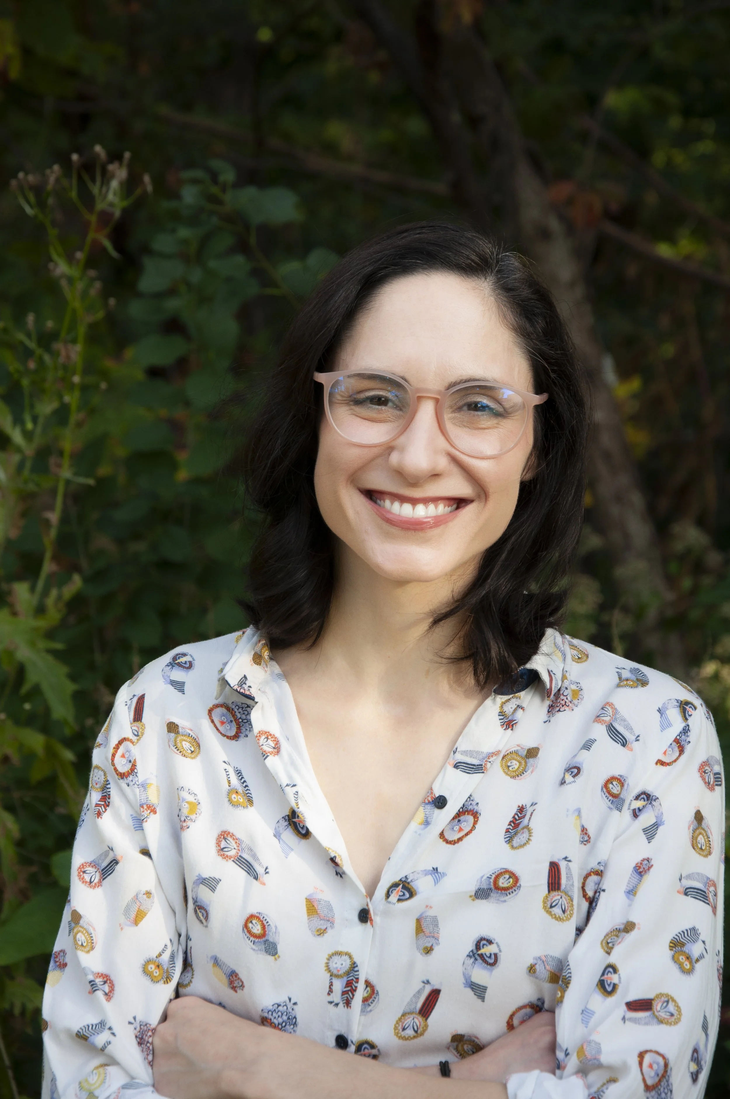 A woman with shoulder-length dark hair, wearing glasses, and smiling with arms crossed, standing outdoors with green leaves and a tree in the background.