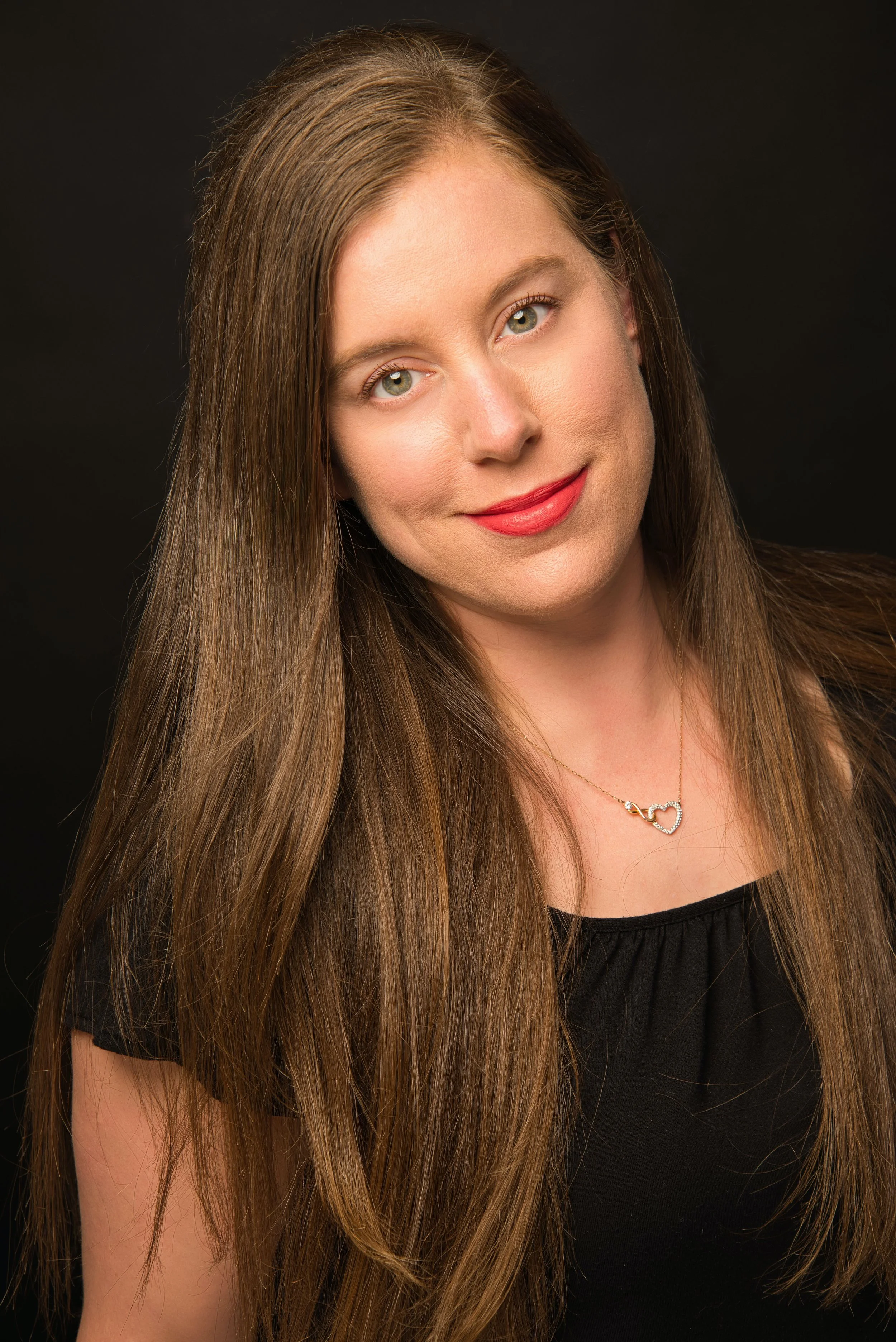 A woman with long brown hair and blue eyes, wearing a black top and a silver heart-shaped necklace, smiling softly against a dark background.