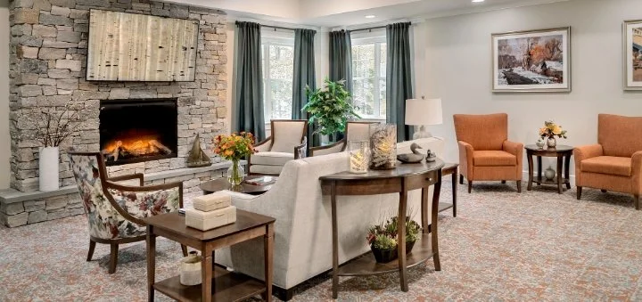 Living room with a stone fireplace, beige and orange armchairs, a gray sofa, a wooden side table, and framed artwork on the wall.