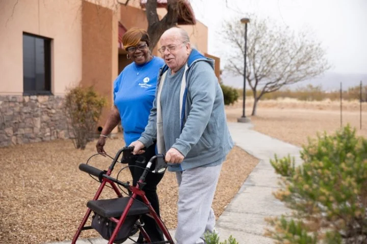 An elderly man using a walker walks along a sidewalk outside a building, with a woman in a blue shirt assisting him and smiling.