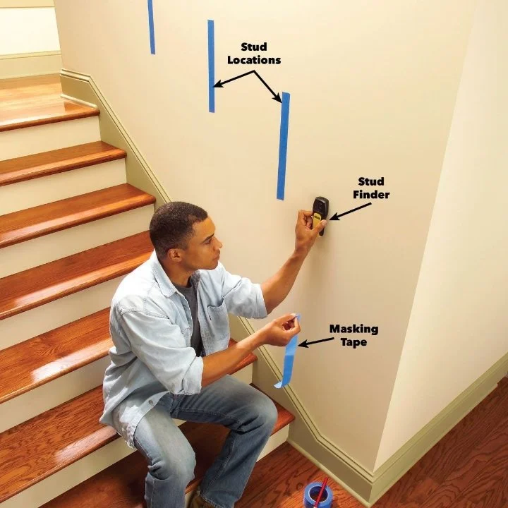 A young man is sitting on wooden stairs applying painter's masking tape to a wall. He uses a stud finder to locate studs behind the wall, indicated by markings and arrows on the wall. The wall has blue tape marking the stud locations, with some tape strips already applied.