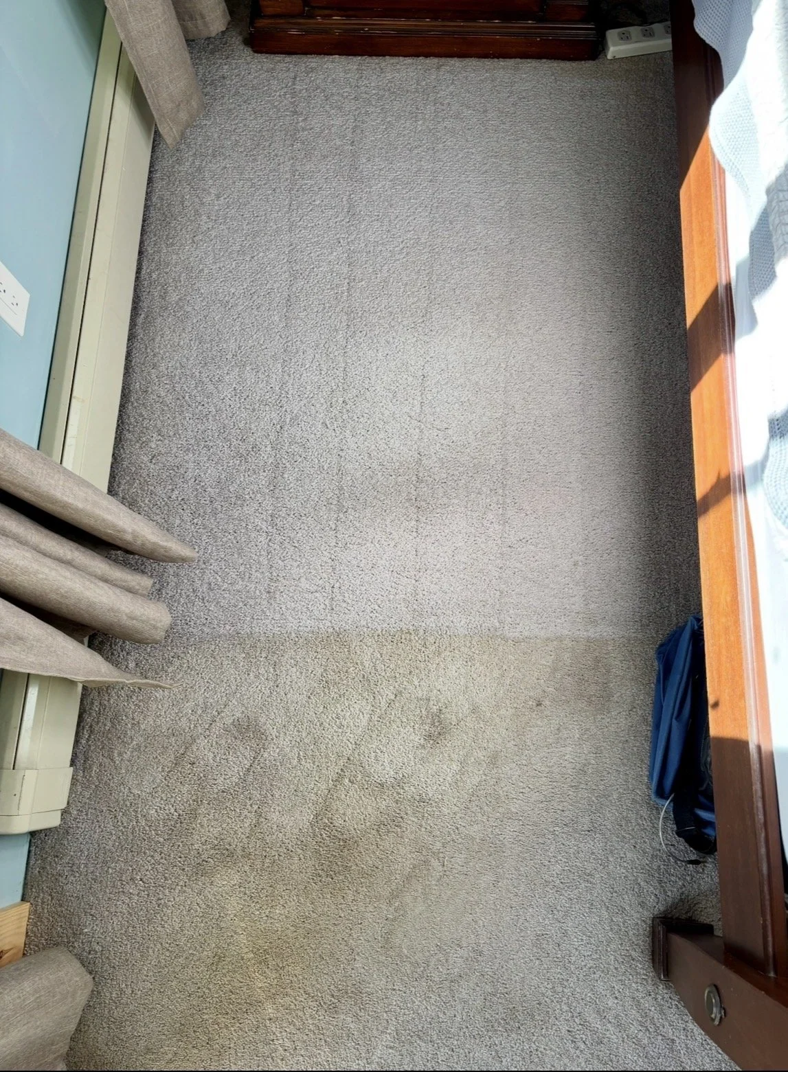 A room with a two-tone carpet; the top part is darker gray, and the bottom part is lighter beige. There's a curtain on the left side, a blue item and electrical outlets near the bottom, and a wooden bed frame or table on the right with a blue backpack underneath.