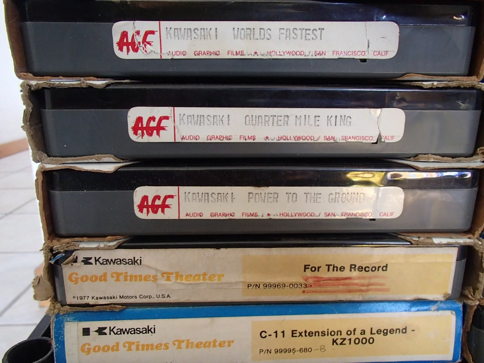 A stack of vintage 8-track tapes with labels featuring titles such as 'Kawasaki World's Fastest,' 'Kawasaki Quarter Mile King,' and 'Kawasaki Power to the Ground.'