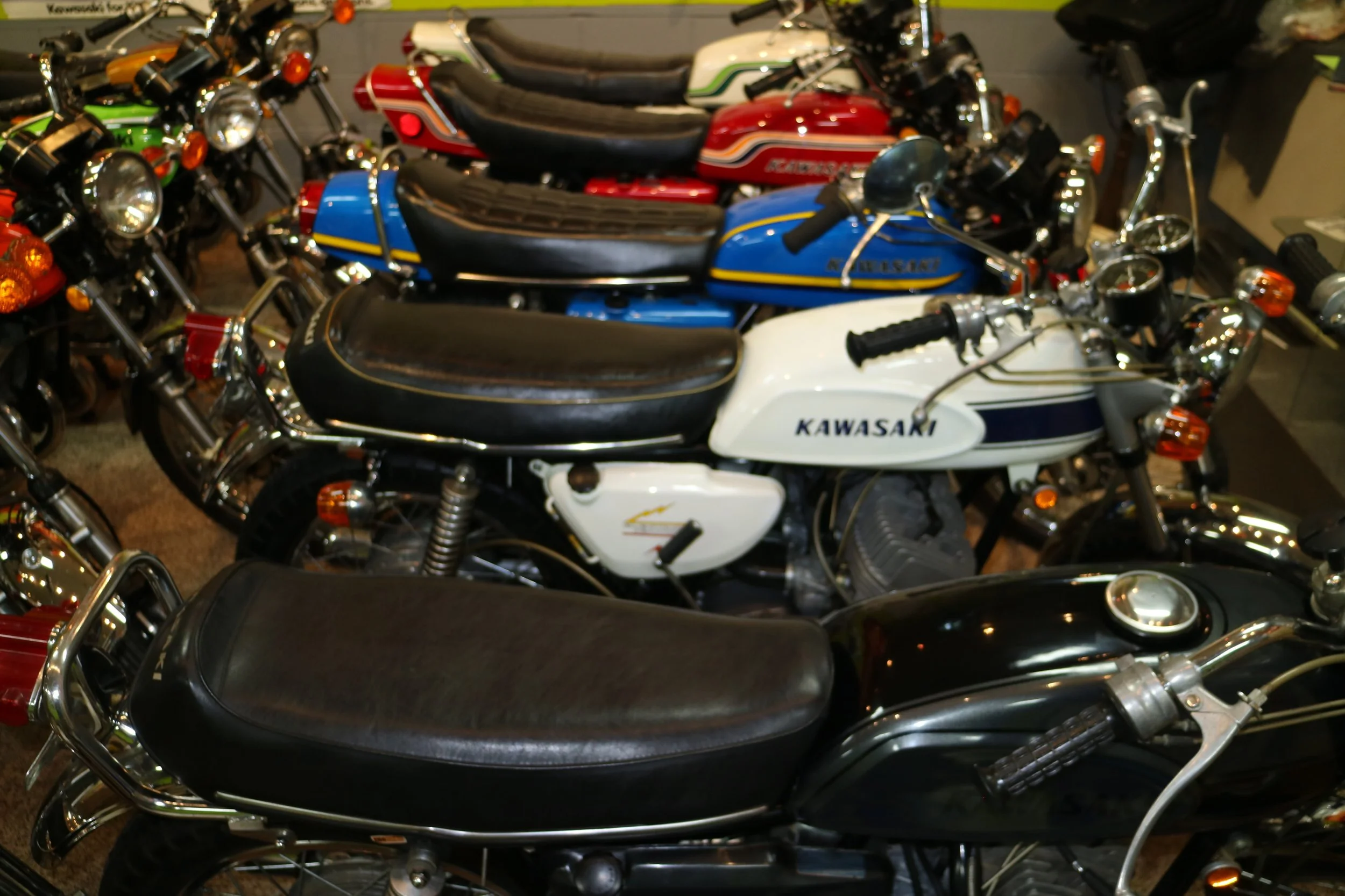 Multiple Kawasaki motorcycles lined up indoors, with various colors including white, blue, red, and green.