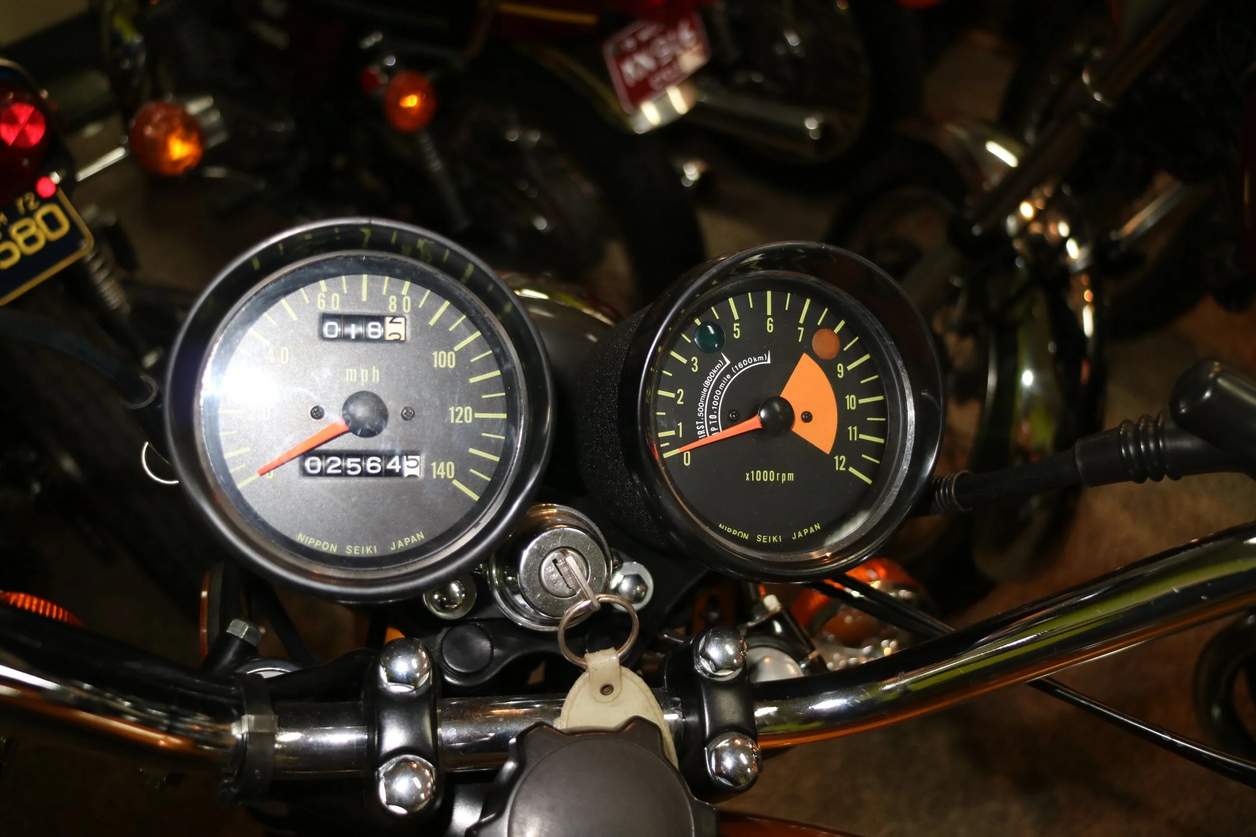 Close-up of motorcycle dashboard with speedometer and tachometer, showing 102,645 miles and engine at around 1,600 RPM, with a background of motorcycle handlebars and other bikes.