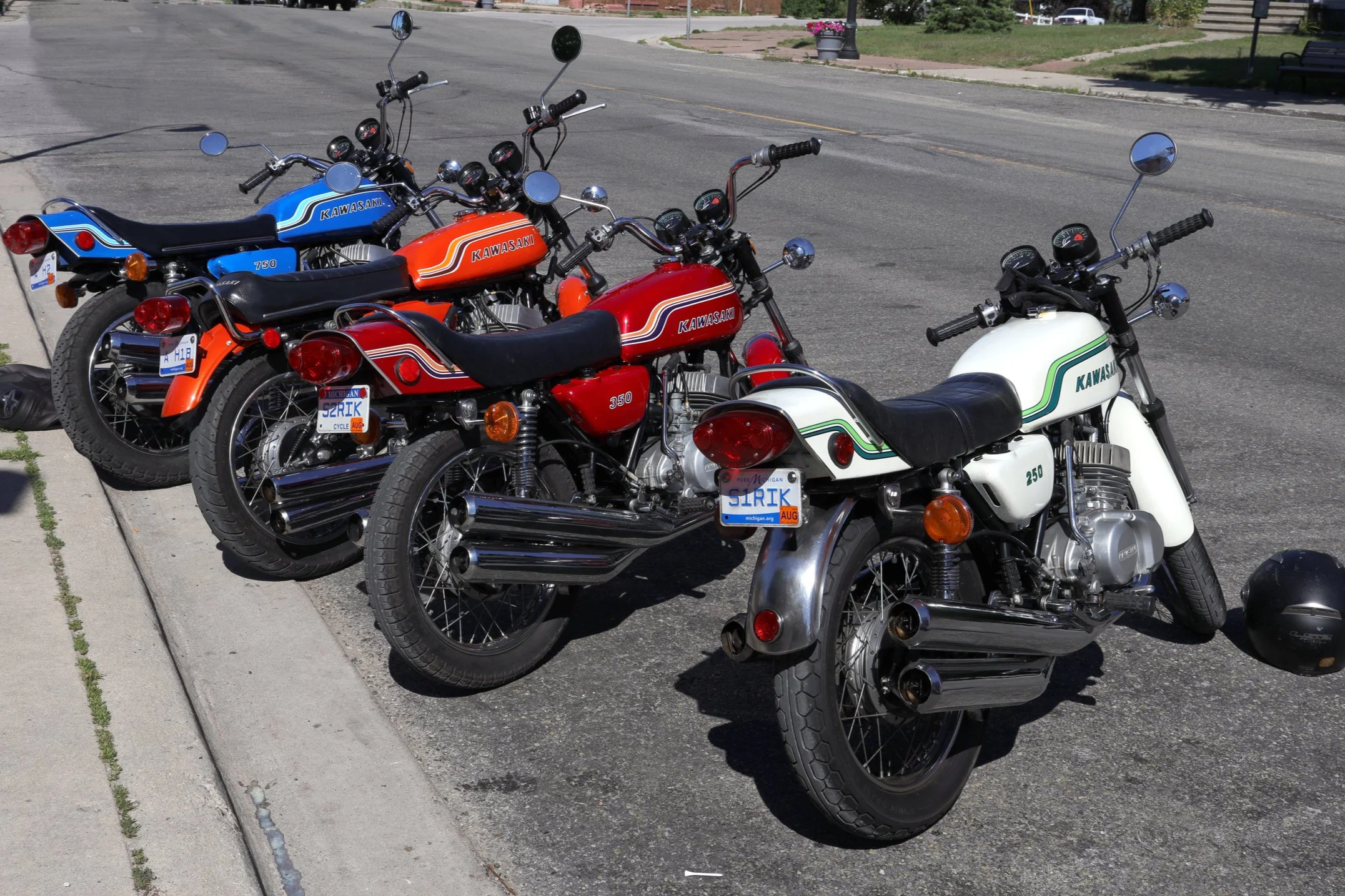 Four Kawasaki motorcycles parked on the side of a street, with different colors: blue, orange, red, and white, in a sunny outdoor setting.