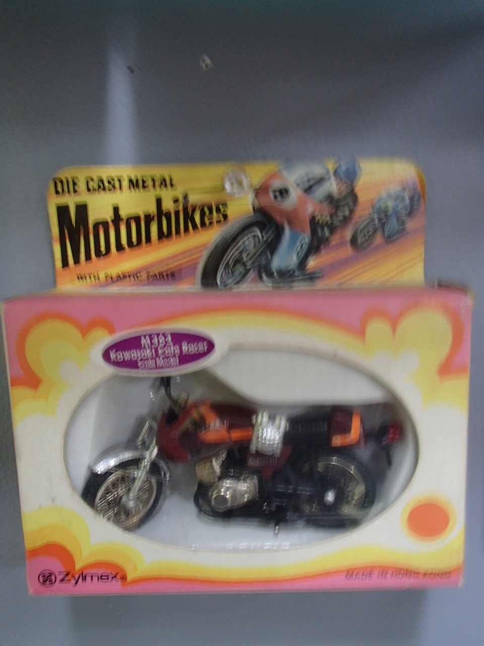 A miniature Kawasaki motorcycle toy inside a pink and yellow box with an oval window, labeled 'Miss Kawasaki Motorcycle Racer,' and a background box with a plastic model motorcycle and rider. The packaging also says 'Die Cast Metal Motorcycle' with '