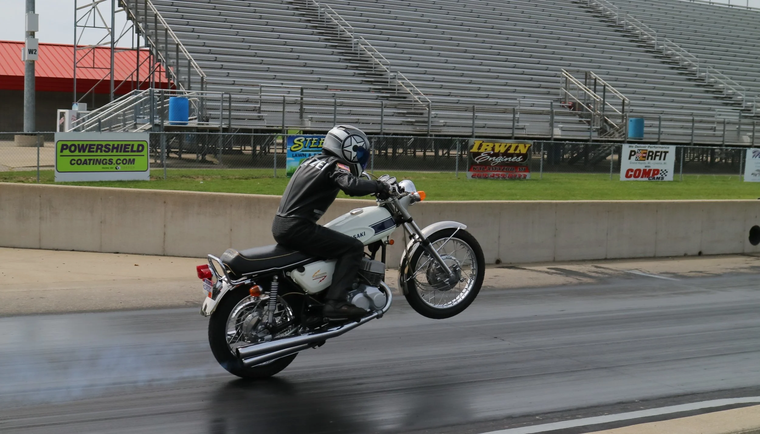 A motorcyclist wearing a helmet and black leather jacket performing a wheelie on a motorcycle on a race track, with empty bleachers and advertising banners in the background.
