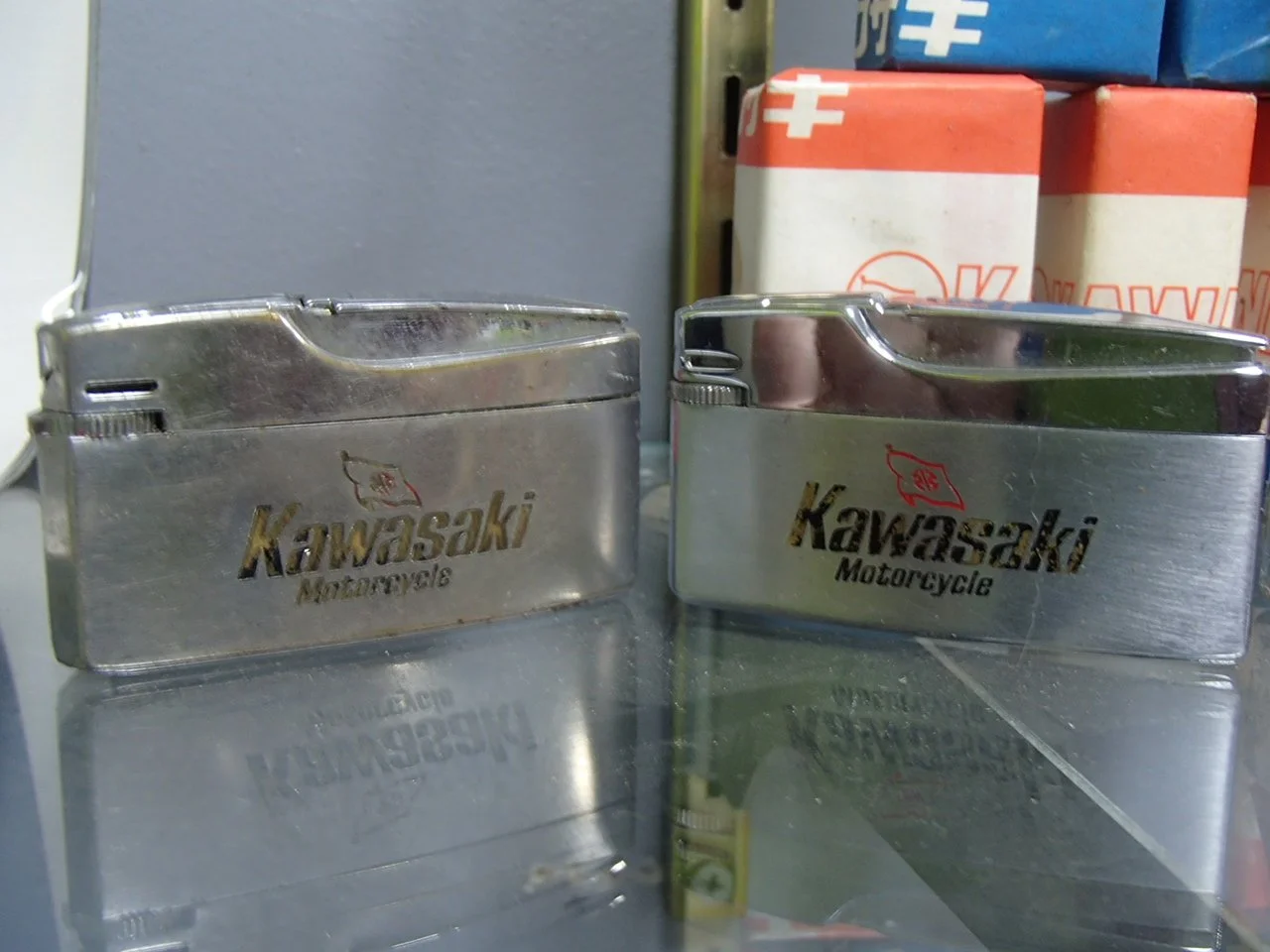 Two vintage Kawasaki Motorcycle metal cigarette lighters with engraved Kawasaki logo, placed on a reflective surface with background boxes.