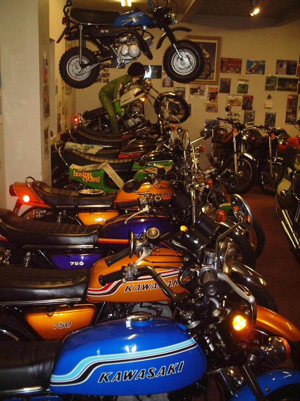 A room filled with vintage Kawasaki motorcycles on display, including models with bright orange, blue, and green paint jobs. There is a motorcycle mounted upside down on the ceiling and a person in a green outfit examining the bikes.