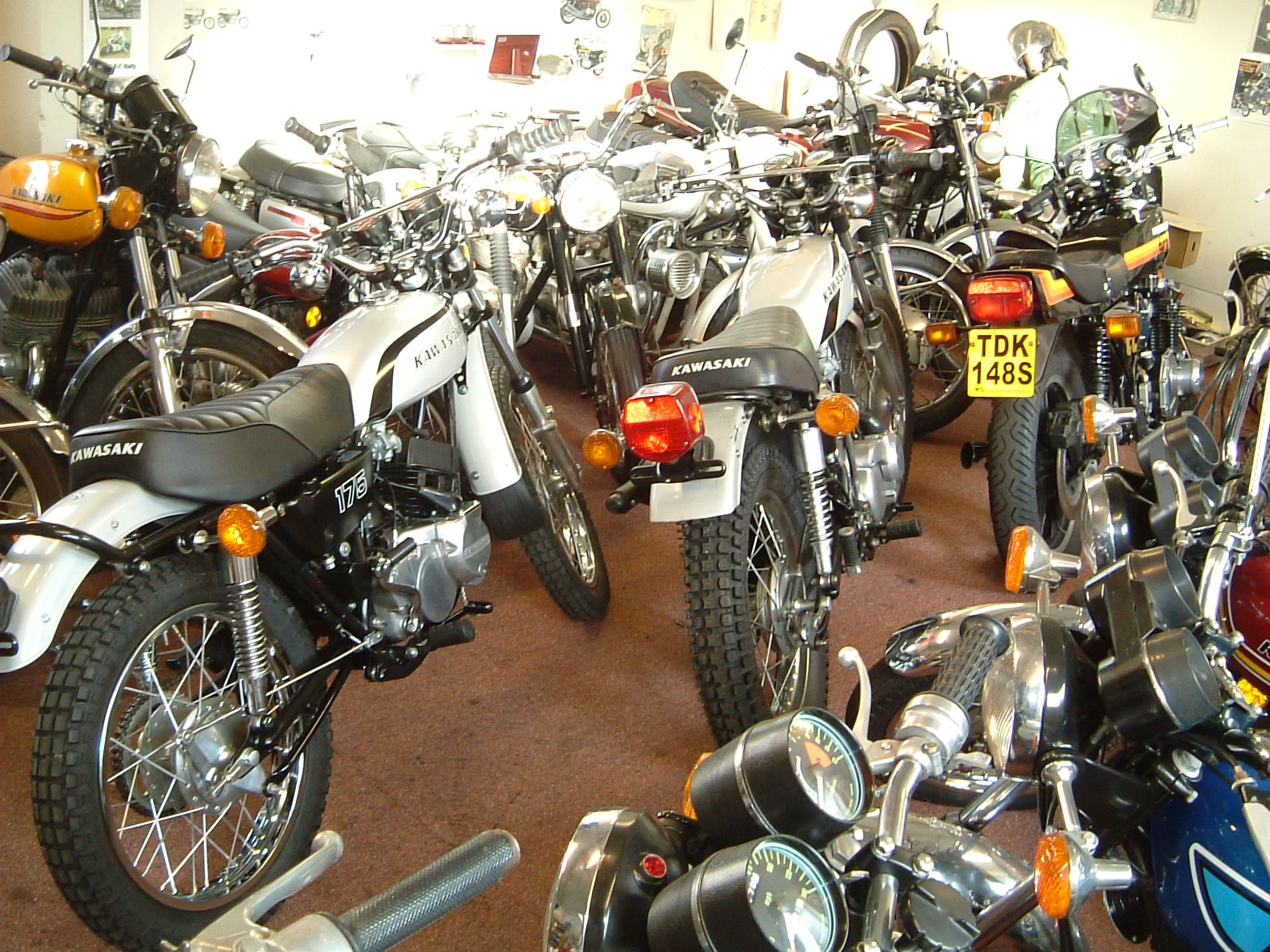 A room filled with numerous vintage motorcycles, some with visible license plates, motorcycles, and a motorcycle helmet.