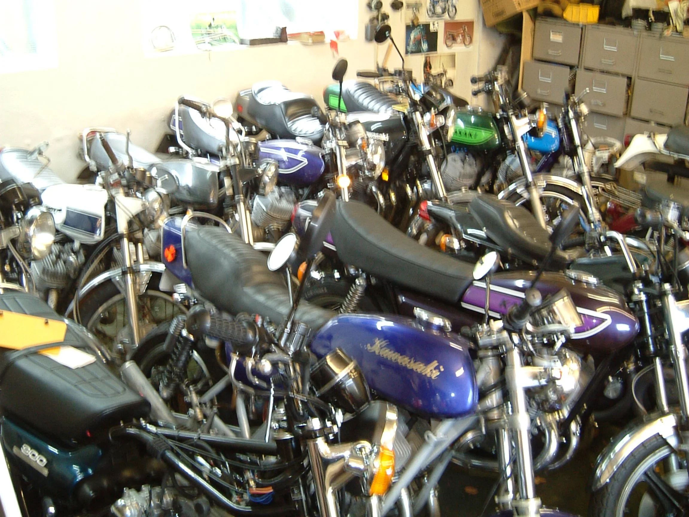 A crowded garage filled with multiple parked motorcycles of various colors, including black, purple, green, and blue, with storage shelves and lockers in the background.