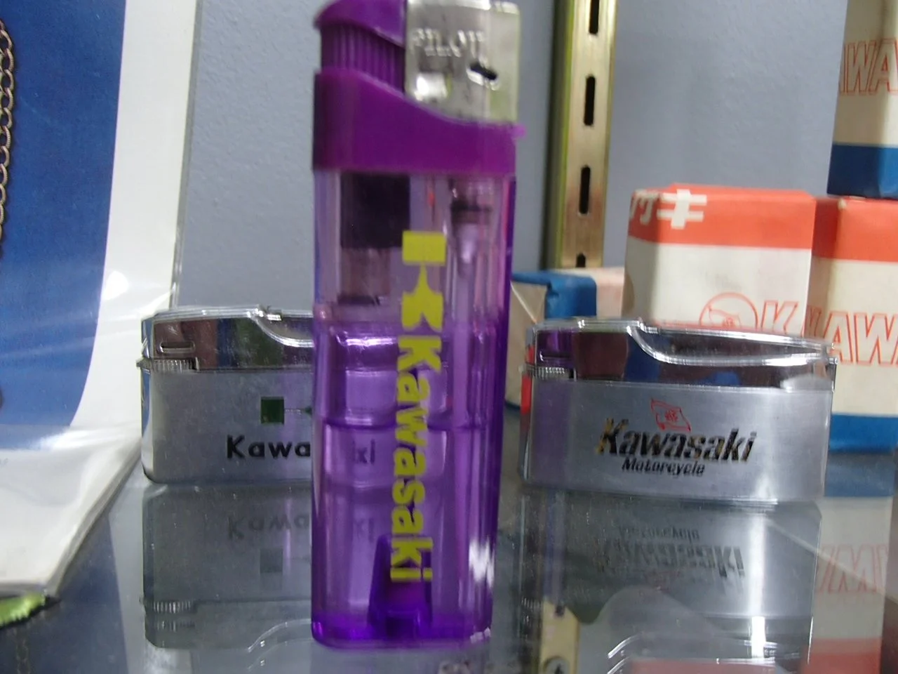 A purple lighter with yellow text standing upright on a reflective surface, with Kawasaki motorcycle parts in the background.