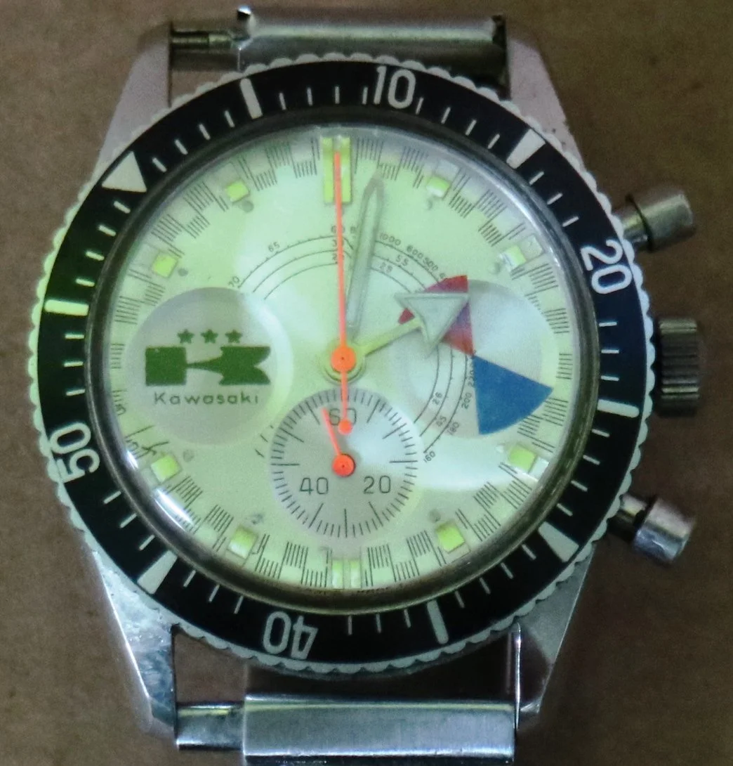 A wristwatch with a yellow face, black bezel, and metal strap. The watch has multiple dials, including a small second hand at the bottom, and features the Kawasaki logo with a green locomotive image on the left side.