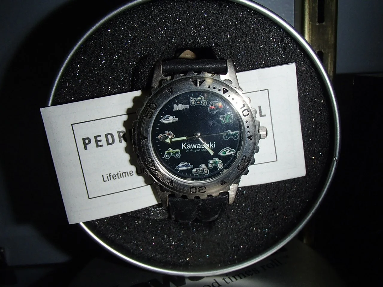 A wristwatch with a black leather strap, featuring motorcycle illustrations on the dial, placed inside a round box with foam padding, resting on a white card with partially visible text, on a black foam surface.