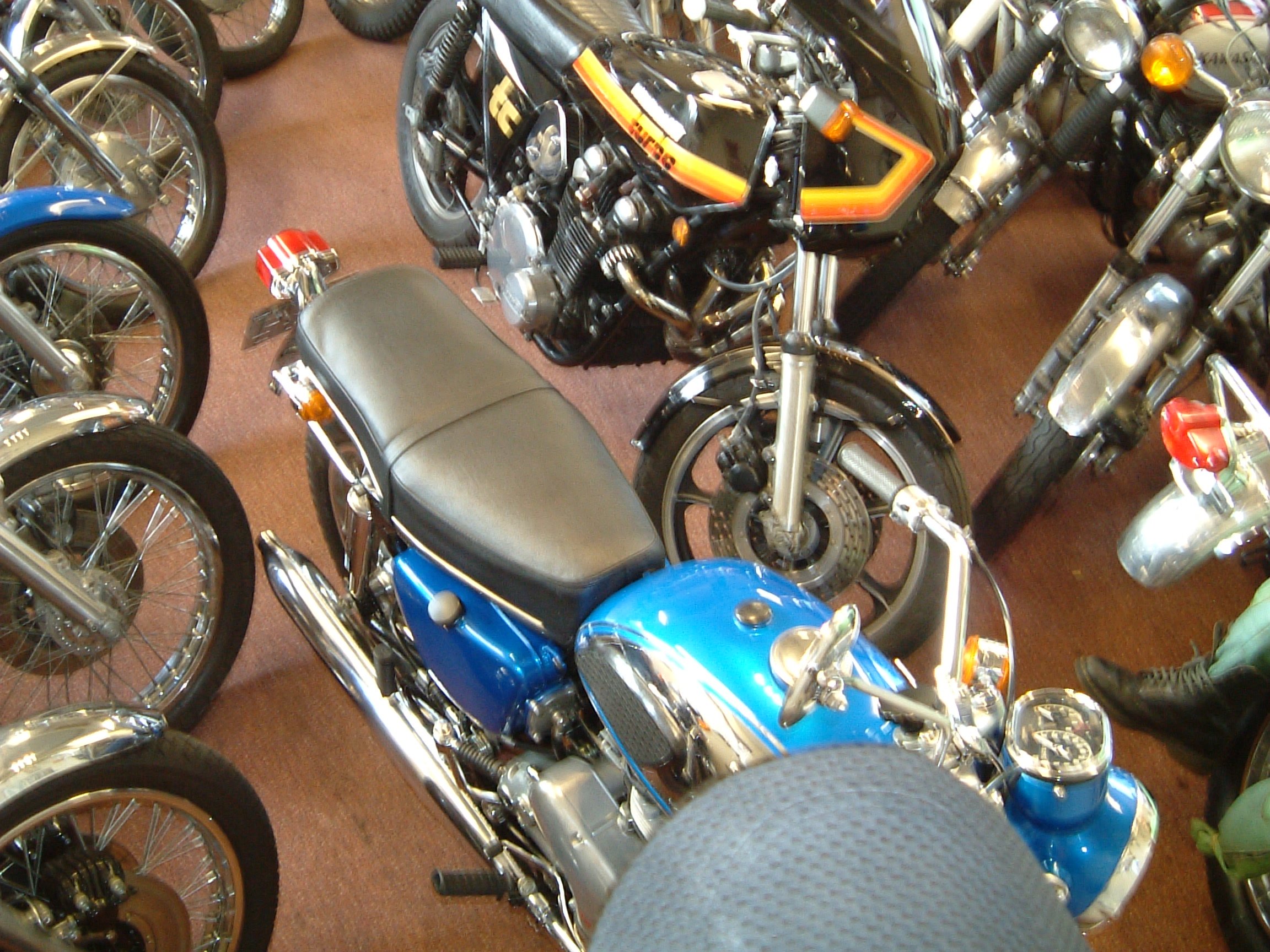 Several motorcycles parked closely together indoors, including a blue motorcycle in the foreground and a black one with orange and yellow detailing in the middle, with other bikes partially visible around them.