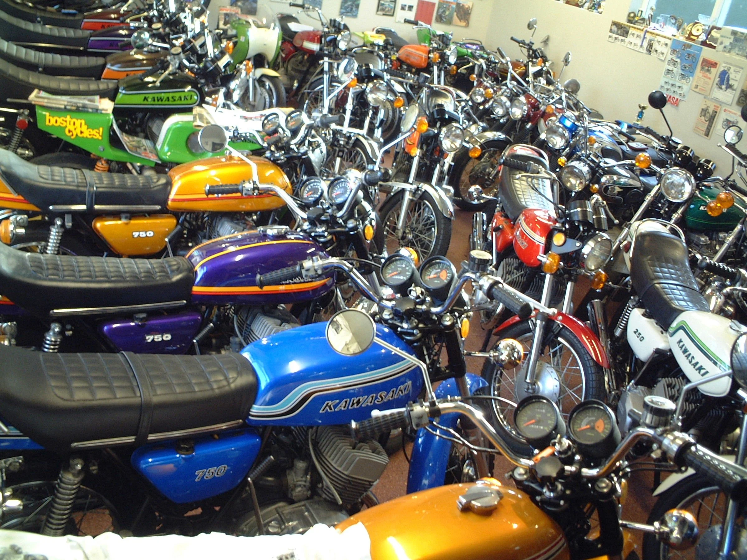 A collection of vintage motorcycles displayed indoors. The motorcycles are closely parked, with visible gauges and prominent manufacturer logos like Kawasaki. The bikes feature bright colors such as green, orange, blue, and black, and have classic st