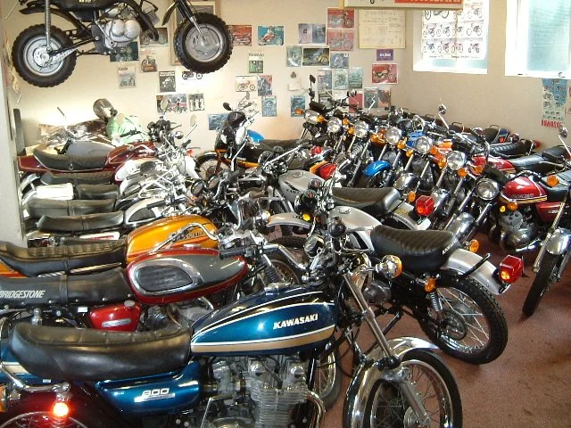 A collection of vintage motorcycles and scooters indoors, with a model motorcycle hanging on the wall and posters in the background.
