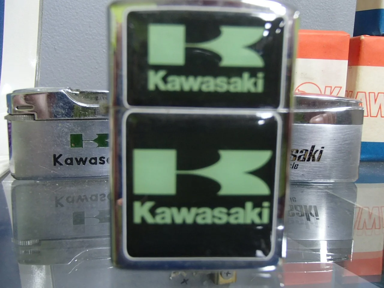 Close-up of a vintage metal lighter with Kawasaki logo, with two similar lighters in the background and some boxes to the right.