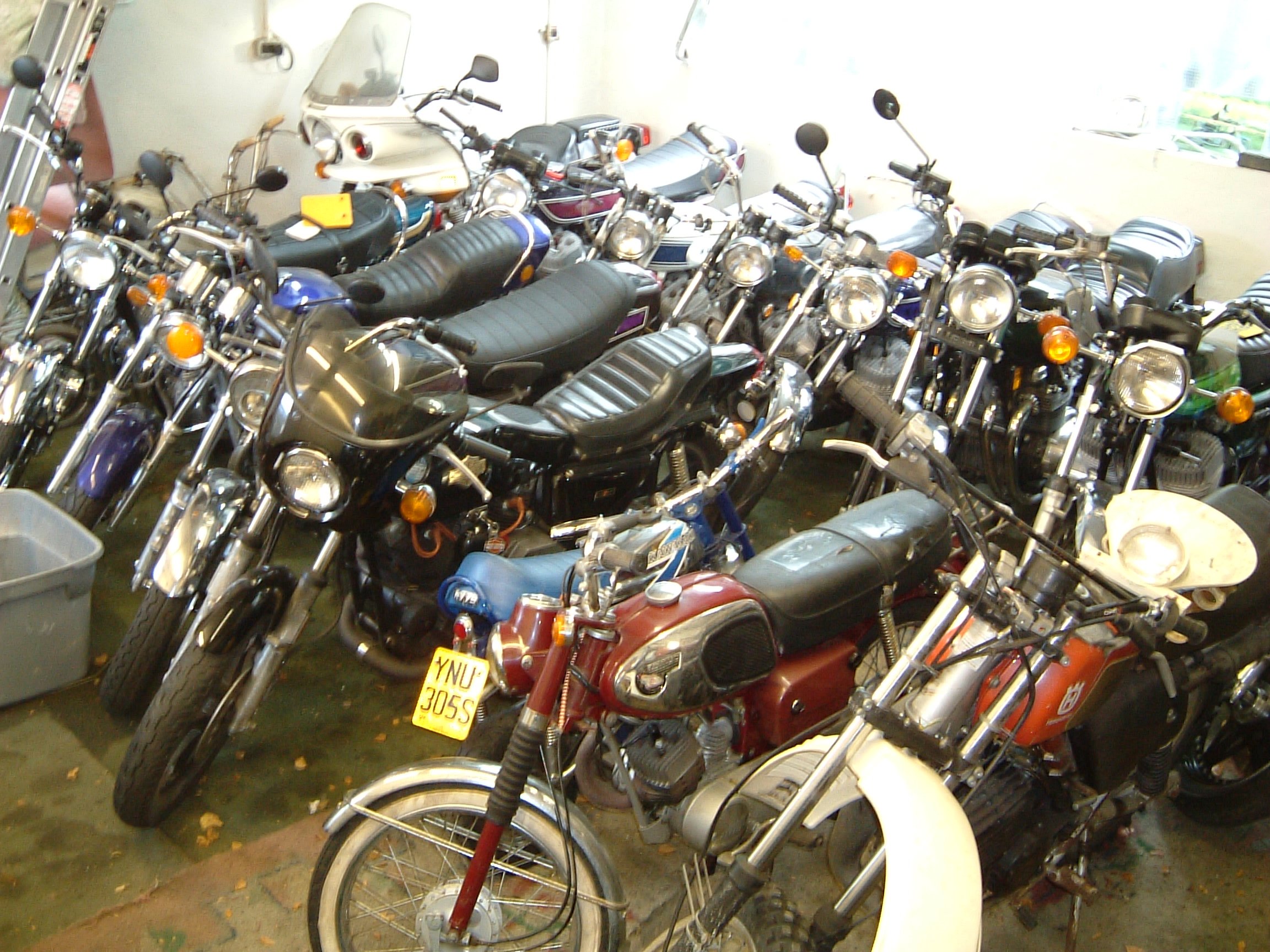 A collection of vintage motorcycles parked indoors alongside a few other vehicles, including a scooter and a car. The motorcycles have different colors and styles, with some featuring black leather seats and chrome accents.