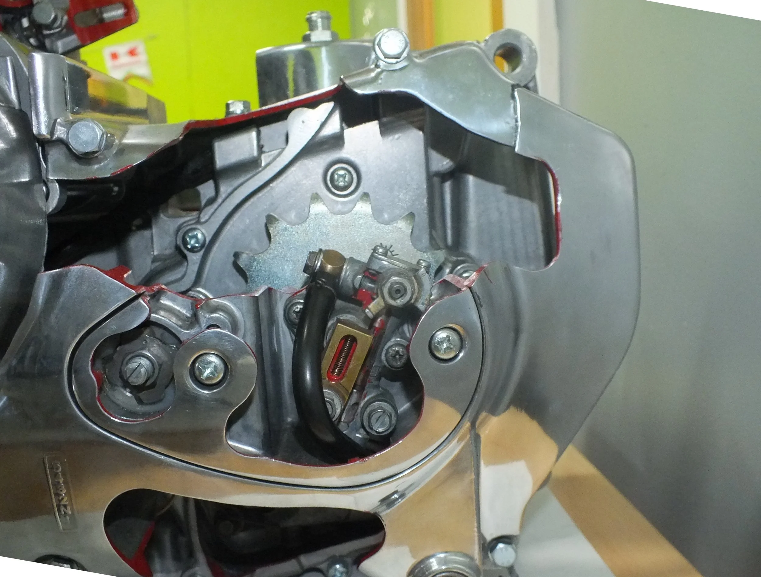 Close-up of a metallic engine part with visible gears, screws, and a red gasket on a workbench, with a green background.