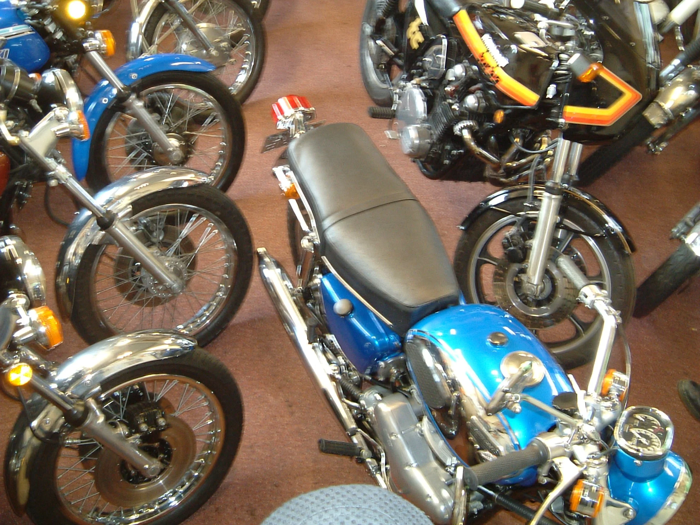 Multiple motorcycles parked indoors, including a blue motorcycle with a black seat in the foreground and another motorcycle with orange detailing and exposed engine components in the background.