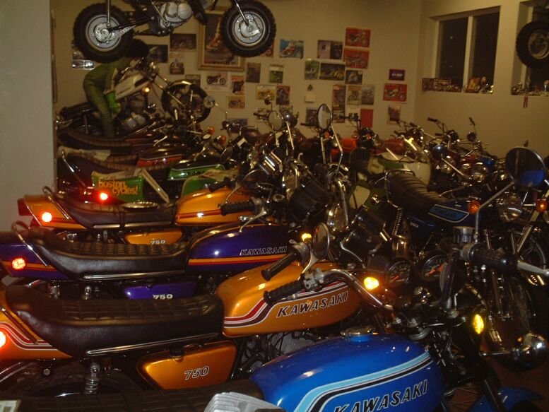 A collection of vintage Kawasaki motorcycles and mopeds in a showroom or garage, with a wall decorated with posters and photos.